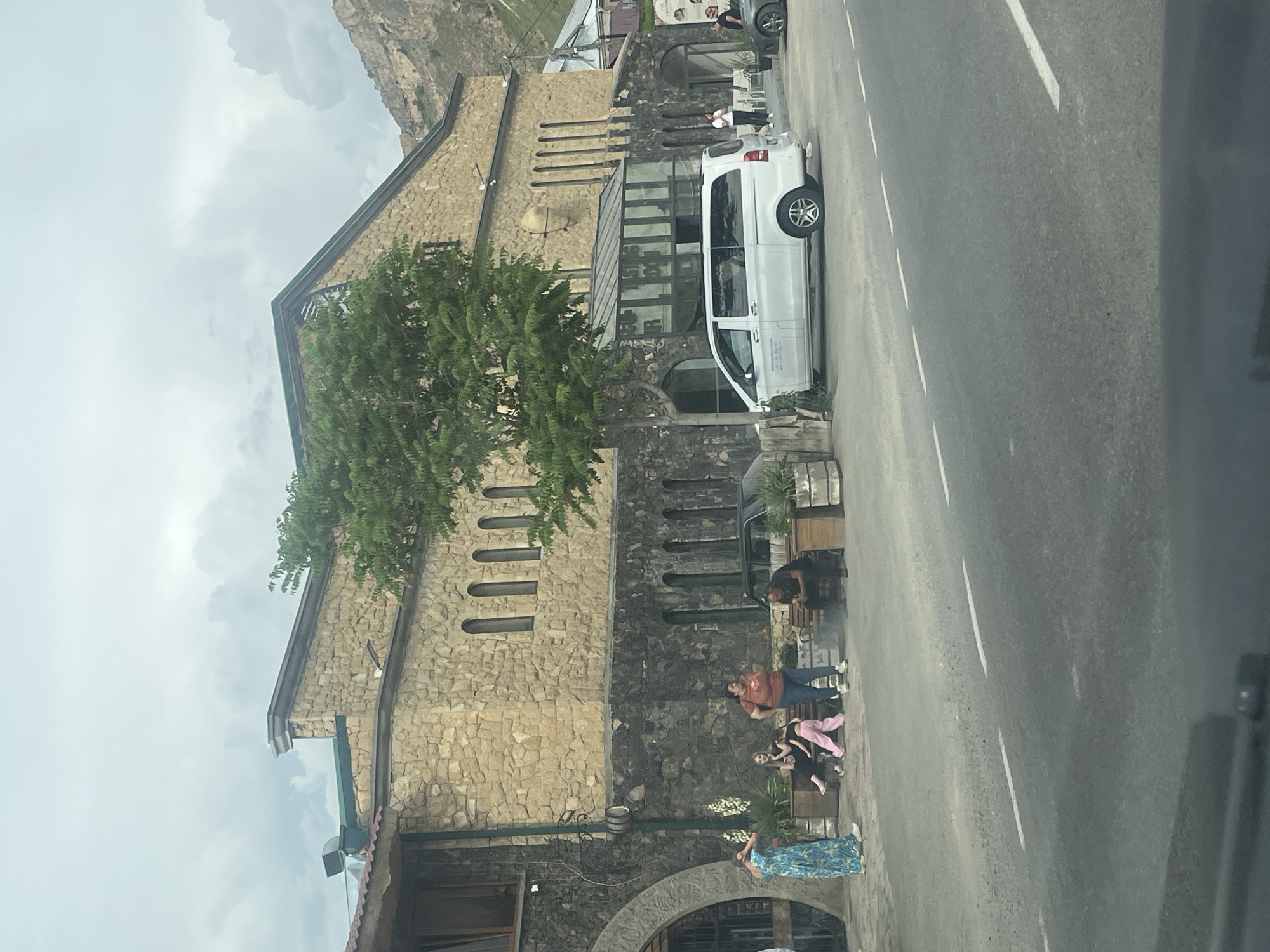 The image, taken in Chiva, Armenia, depicts a rustic stone building adjacent to a paved road, set against a backdrop of mountains and a cloudy sky. The building is constructed with varied stone masonry in shades of light and dark brown, featuring a slanted dark roof and several arched windows and entrances. A prominent green-leafed tree stands in front of the building.
A group of people are gathered outside the building on the left side of the road. One woman, wearing a long blue and white patterned dress, stands near the foreground. Further back, a man in a reddish-brown shirt and blue jeans is standing, while others are seated on a bench or low wall, including one person in pink pants. They appear to be socializing or relaxing outdoors.
A white Mercedes-Benz van is parked on the side of the road near the building, with a dark SUV partially visible further down the road. Wooden barrels are placed near the building's facade, possibly for decoration or practical use. The road itself has white dashed lines. The scene suggests a daytime setting under an overcast sky. No legible text is visible on signs or vehicles.