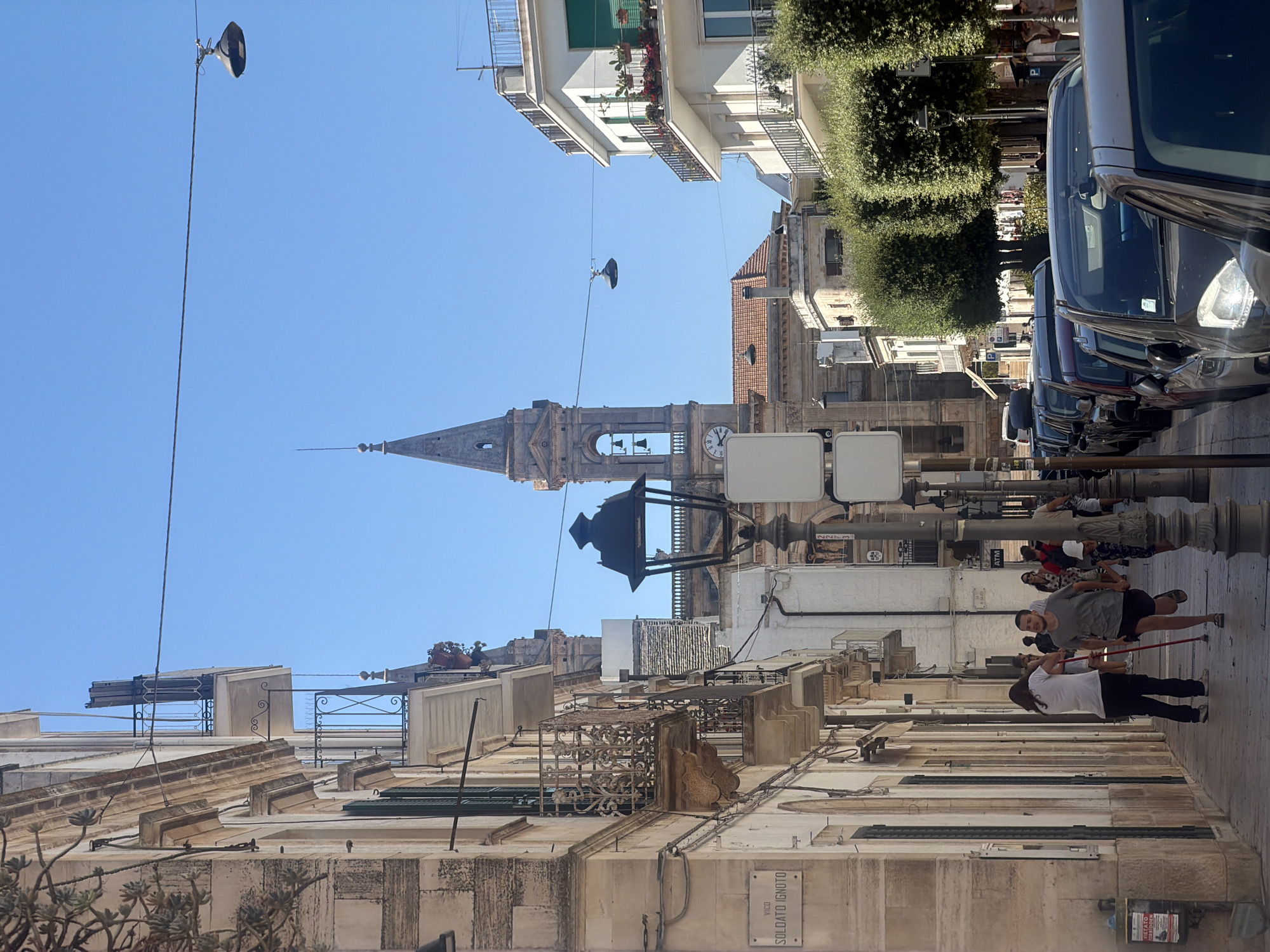 This image depicts a street scene in Alberobello, Italy, under a clear blue sky. The left side of the frame features a building with a light-colored facade and ornate balconies. A street sign reading "VICO SOLDATO IGNTO" is visible. On the right side, a white building with balconies and potted plants is situated. A prominent bell tower with a clock face rises in the background, partially obscured by other buildings and trees. The street is lined with parked cars. In the foreground, several people are walking along the sidewalk. One person in a white t-shirt and dark pants walks away from the camera, while another in a grey t-shirt and shorts runs towards the camera, holding a red pole. Other individuals are visible in the background. A street lamp with an ornate design stands prominently in the middle ground. The overall impression is that of a sunny day in a historic Italian town.