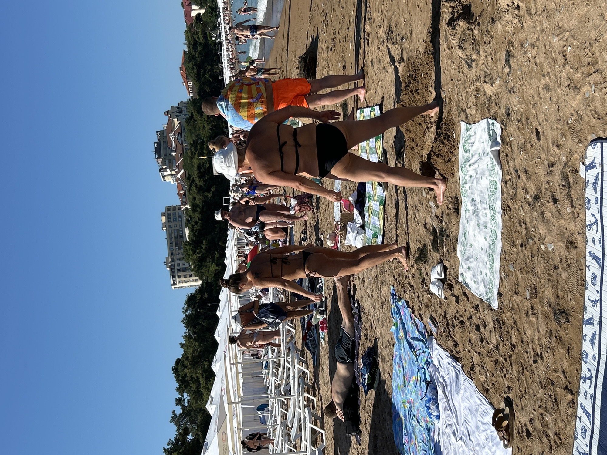 The image depicts a crowded beach on a sunny day, with numerous people engaged in various activities. In the foreground, several individuals are sunbathing and relaxing on towels and beach chairs. A man is lying on his stomach on a towel, facing away from the camera. A woman in a black bikini stands in the center, facing the water. Another woman, also in a bikini, is standing to her left, holding a towel. Further back, more people are visible walking along the beach or in the shallow water. The background shows a line of white beach umbrellas, a lush green tree line, and several multi-story buildings, suggesting an urban coastal setting. The buildings and the overall landscape are consistent with the architecture and geography found in Gelendzhik, Russia, a popular resort city on the Black Sea coast. The scene is typical of a busy summer day at a beach resort.