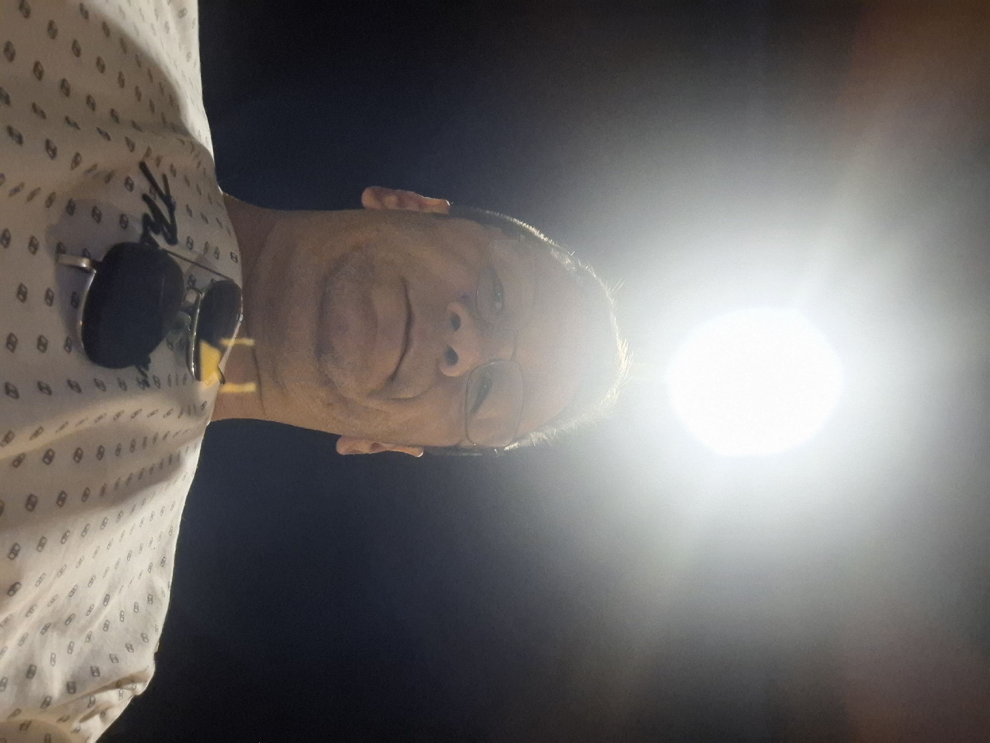 The image is a low-angle, selfie-style shot of a man wearing glasses and a light-colored, patterned shirt. He is facing upwards towards a bright, circular light source that is causing lens flare and washing out the upper portion of the image. The man's face is partially obscured by shadow and the bright light. A pair of sunglasses is resting on his shirt. The background is dark and appears to be a night sky. The pattern on his shirt is a repeating geometric design. There is no identifiable information in the image that would suggest a specific location within the city of Yerocham, Israel.