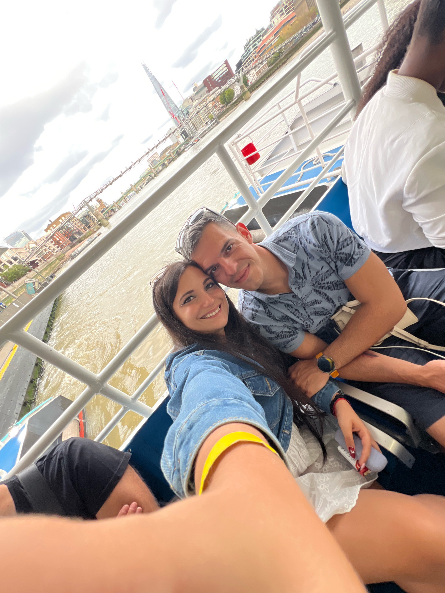 The image shows a couple taking a selfie on a boat tour on the River Thames in London. In the background, several London landmarks are visible, including the Shard and Tower Bridge. The couple appears to be enjoying their time together, with both smiling at the camera. The water in the river is murky brown, indicating it is a cloudy day. The boat they are on has white railings and blue seating.