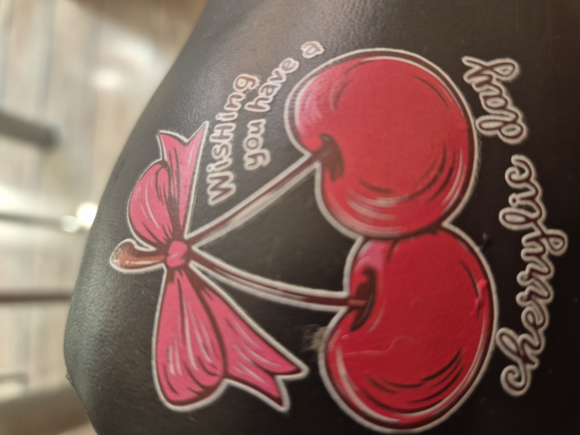 A sticker featuring two red cherries tied with a pink bow is adhered to a dark, textured surface, likely a bike seat or bag. The sticker also contains white text that reads, "Wishing you have a cherrylic day". The background is blurred and out of focus, suggesting an indoor or shaded outdoor environment.