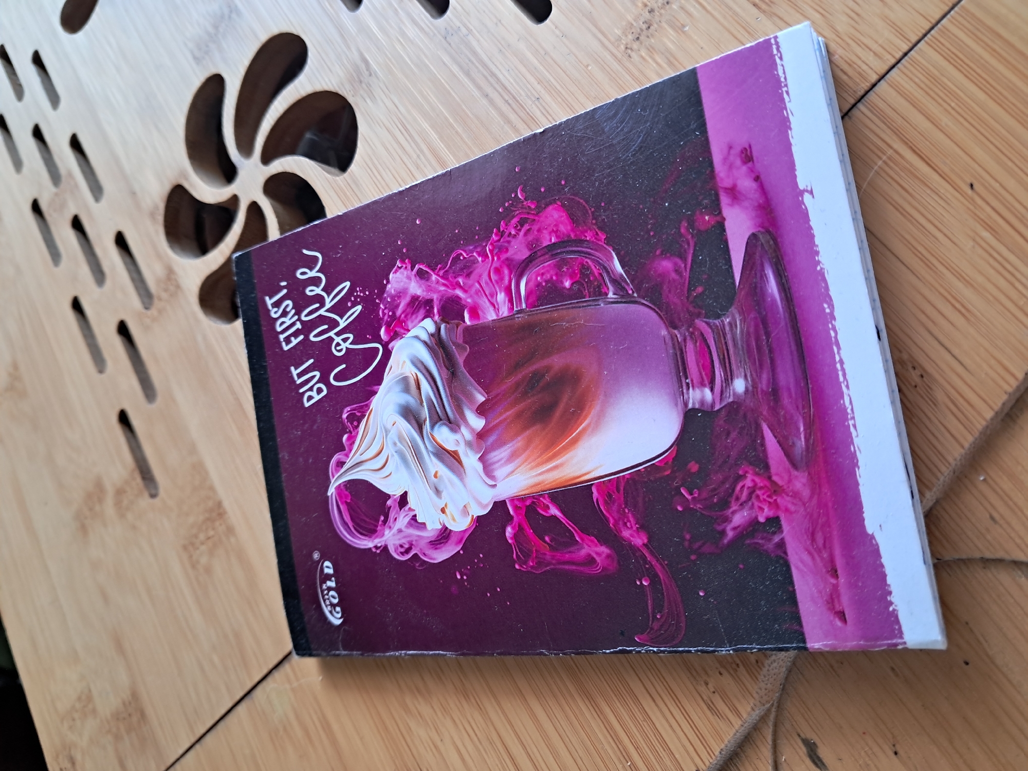 This media file presents a small, vertically oriented notebook or diary resting on a light-colored wooden surface in Kyiv, Ukraine. The notebook cover features a vibrant, artistic illustration: a clear glass or mug, possibly containing coffee, topped with a rich swirl of white and brown-orange frothy cream or whipped topping. Surrounding the glass and extending across the dark background are dynamic, swirling patterns of magenta and pink liquid or smoke-like elements.
Visible text on the cover includes the phrase "BUT FIRST, Coffee" written in a stylized, cursive font, and a small logo near the bottom left featuring the word "GOLD."
The wooden surface beneath the notebook is characterized by intricate carved patterns, specifically several rows of short, parallel slits and a larger, abstract floral or pinwheel-shaped cut-out. These openings reveal a darker material underneath, suggesting the wooden piece might be a decorative table, a slatted tray, or a stand.
The image is captured during the daytime, indicated by the bright, natural-looking light and shadows. The scene is static, depicting no active event or people, focusing solely on the aesthetically pleasing composition of the notebook and its immediate surroundings.