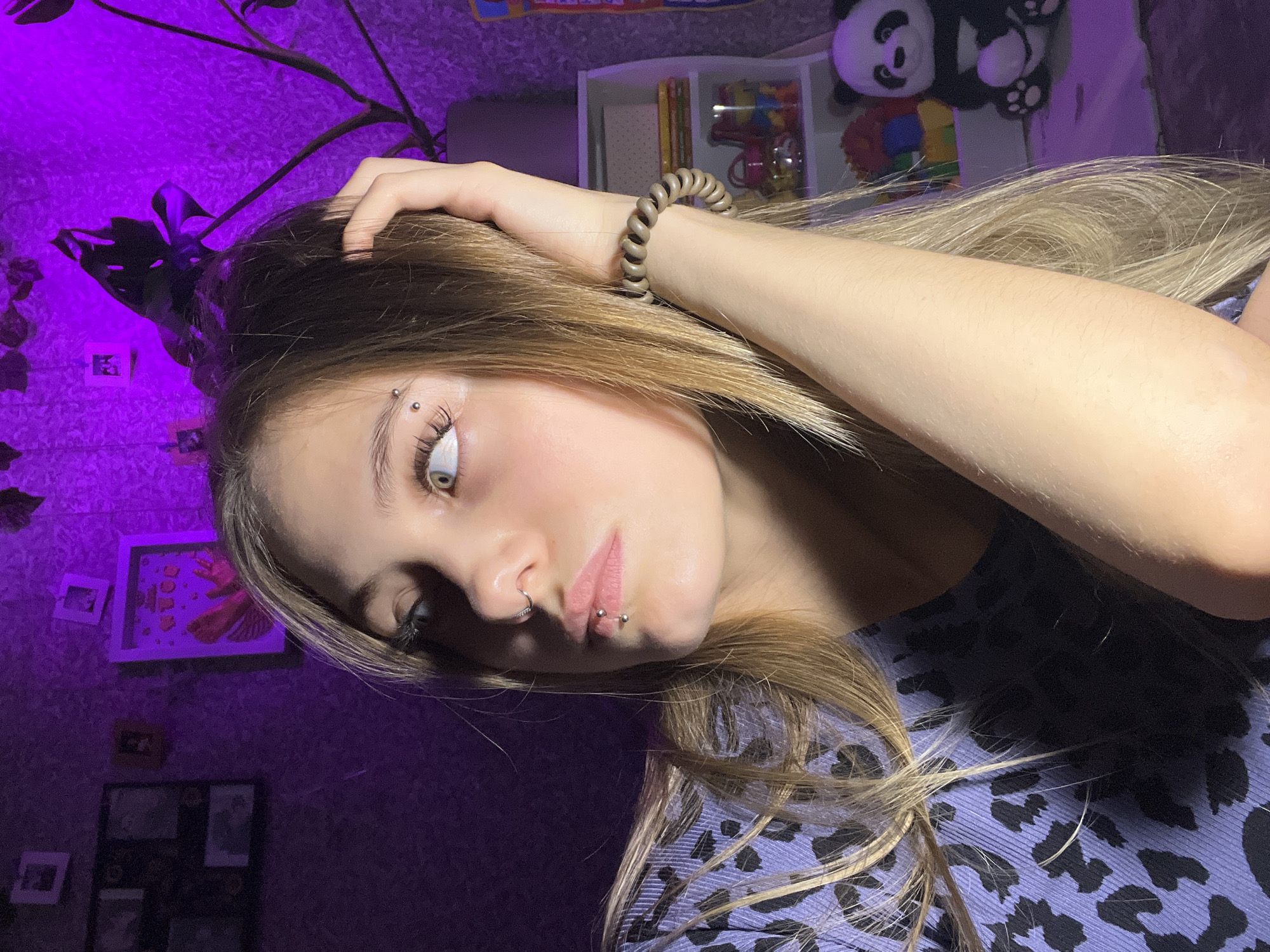 A selfie shows a young woman with light brown hair, possibly lightened, resting her hand on her head. She has an eyebrow piercing on her left eyebrow and a lip ring. She wears a brown, coiled hair tie on her wrist. She is wearing a blue and black animal print top. The background shows a purple-lit wall with framed pictures, a plant, and a shelf with toys, including a panda plush. The location Фоки, Russia is not indicated in the image.