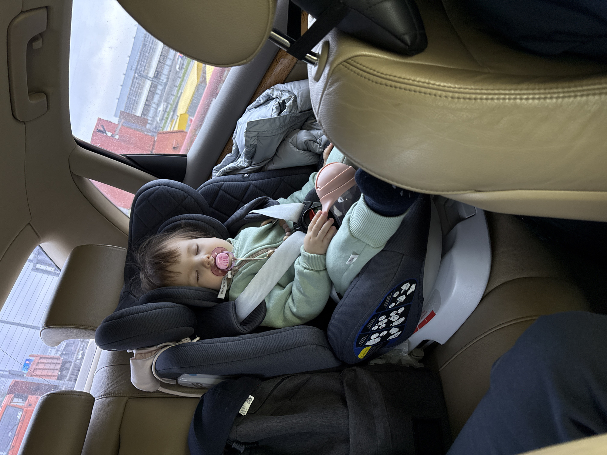 A young child sleeps in a car seat. The child is wearing a light green sweatshirt and has a pacifier in their mouth. The car seat is in the back of a car with beige leather interior. The car is in Moscow, Russia, as evidenced by the view out the window of an industrial area with red brick buildings and metal structures. The weather appears to be overcast. There is a grey jacket or blanket behind the child. A black bag is under the car seat. A leg in dark blue fabric is seen next to the seat, likely belonging to another passenger.