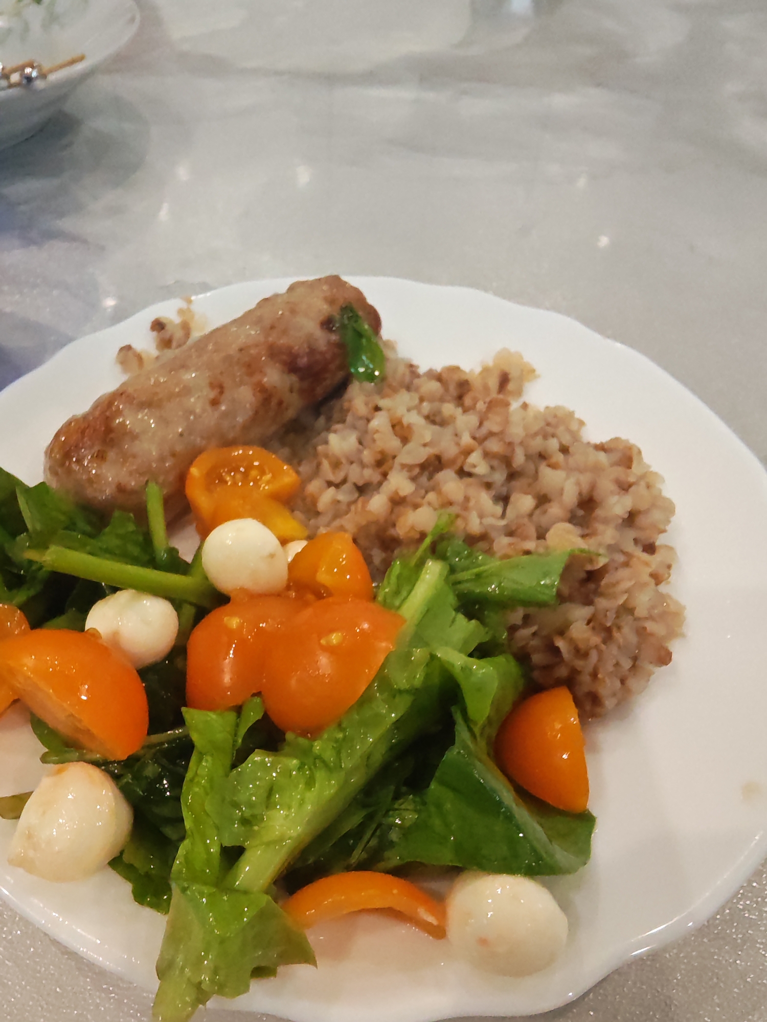 A close-up shot captures a meal presented on a white, slightly irregularly shaped plate. The dish consists of three main components: a meat item, buckwheat, and a fresh salad.
On the left side of the plate is a large, light brown meat item, resembling a grilled or baked sausage or a meat roll, with some browning on its surface and a few green flecks. To its right, covering the upper portion of the plate, is a generous serving of cooked buckwheat (kasha), characterized by its light to medium brown grains.
The lower half of the plate is occupied by a colorful salad. It features vibrant green leafy vegetables, likely lettuce or spinach, mixed with several halved orange cherry tomatoes. Interspersed among the greens and tomatoes are small, spherical white pieces, strongly resembling mozzarella pearls. The salad ingredients appear fresh and slightly glistening, suggesting a light dressing.
The background is a blurry, light-colored surface, possibly a table, showing subtle reflections. A small portion of another out-of-focus white plate is visible in the upper left corner, with some indistinct, thin objects on it. The scene suggests an indoor dining setting. No people or visible text are present in the image.