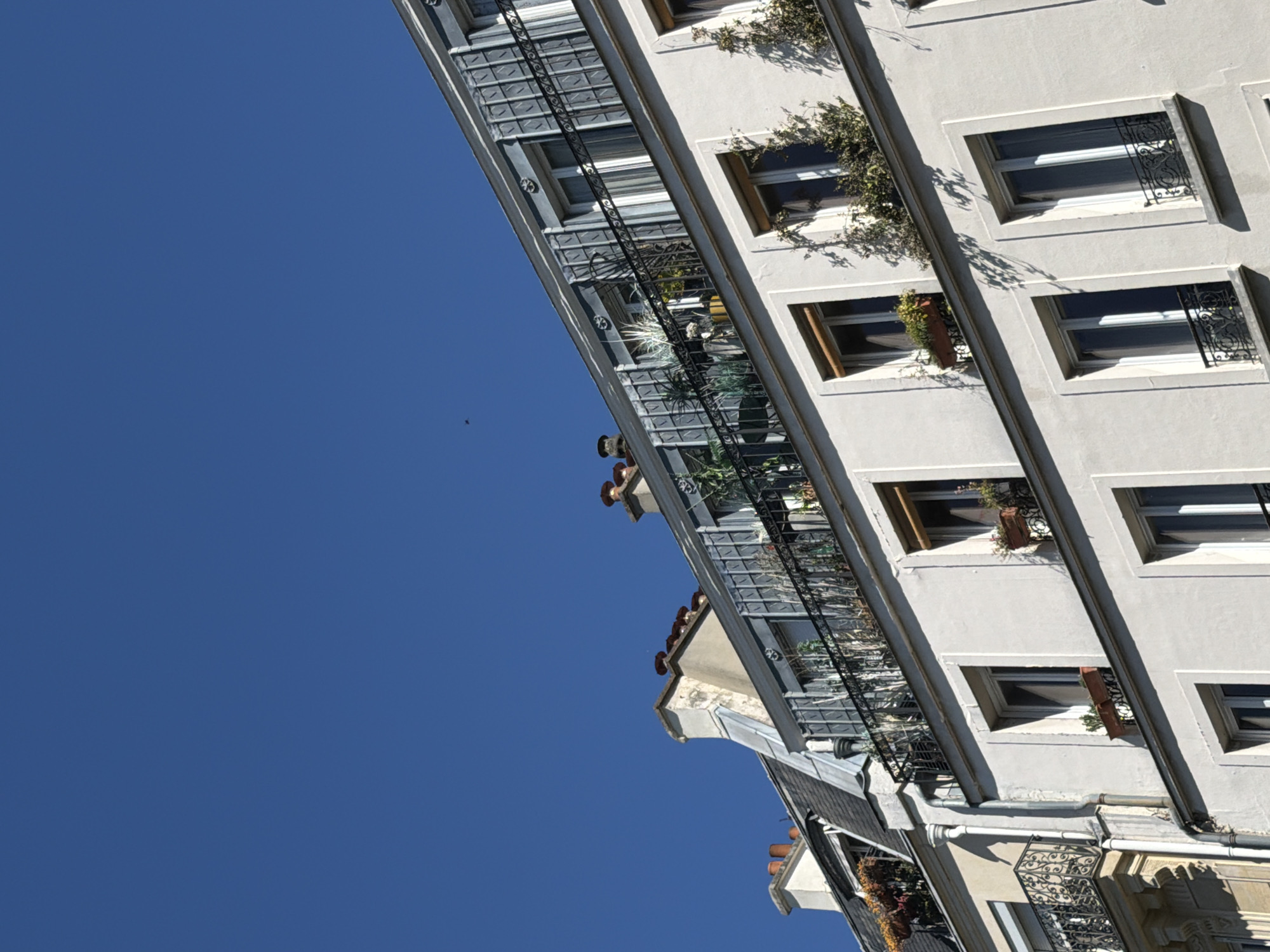 The image is a low-angle shot of a building in Paris, France, under a clear blue sky. The building's facade is light-colored, possibly white or gray, and it features rows of windows with black metal grilles. Balconies are adorned with potted plants and greenery. Two people are visible on one of the balconies. The architectural style is typical of Parisian buildings, with intricate details and classic lines.