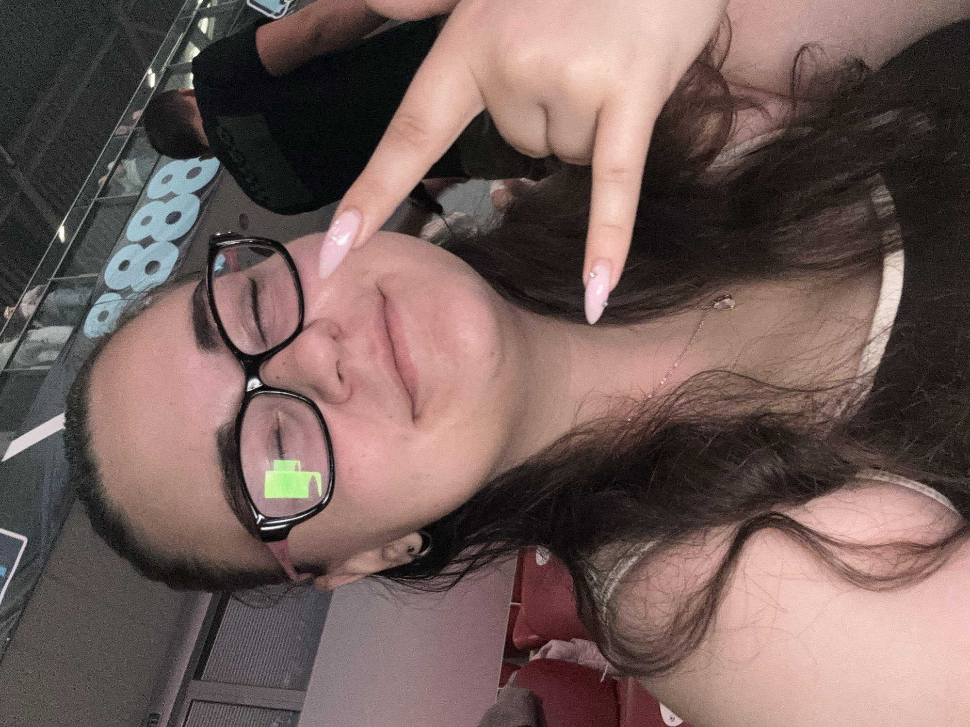 A young woman poses for a selfie indoors. She is seated in a stadium or theater, as indicated by the presence of red seats behind her. She wears glasses with a green sticker on one lens and has long, dark brown hair. Her nails are manicured with a light pink polish and rhinestones. She is making a hand gesture, pointing downward with her index and pinky fingers extended. There is another person in the background, wearing a black shirt. The numbers "888" are visible in the background, possibly indicating a brand or logo. The location is likely in Sofia, Bulgaria, as the "888" logo is associated with 8888.bg, a Bulgarian betting and gambling website with sports sponsorships at venues in Sofia.