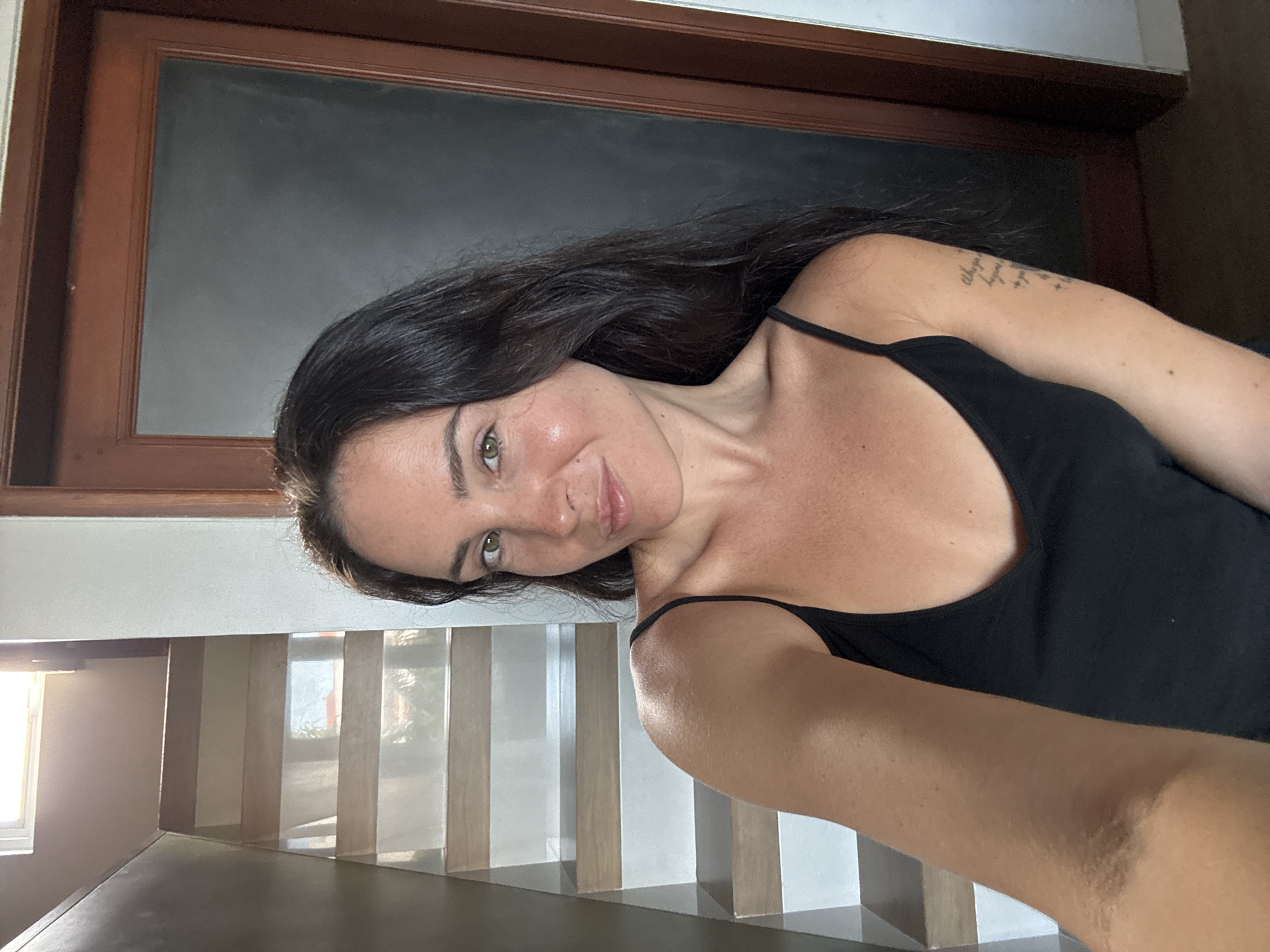 In North Kuta, Indonesia, a woman with long, dark hair and green eyes takes a selfie. She is wearing a black tank top. Behind her, a modern staircase with wooden steps and white risers is visible, illuminated by natural light from a window at the top. To her right, a dark-paneled door with a wooden frame is present. A tattoo is visible on her left arm.