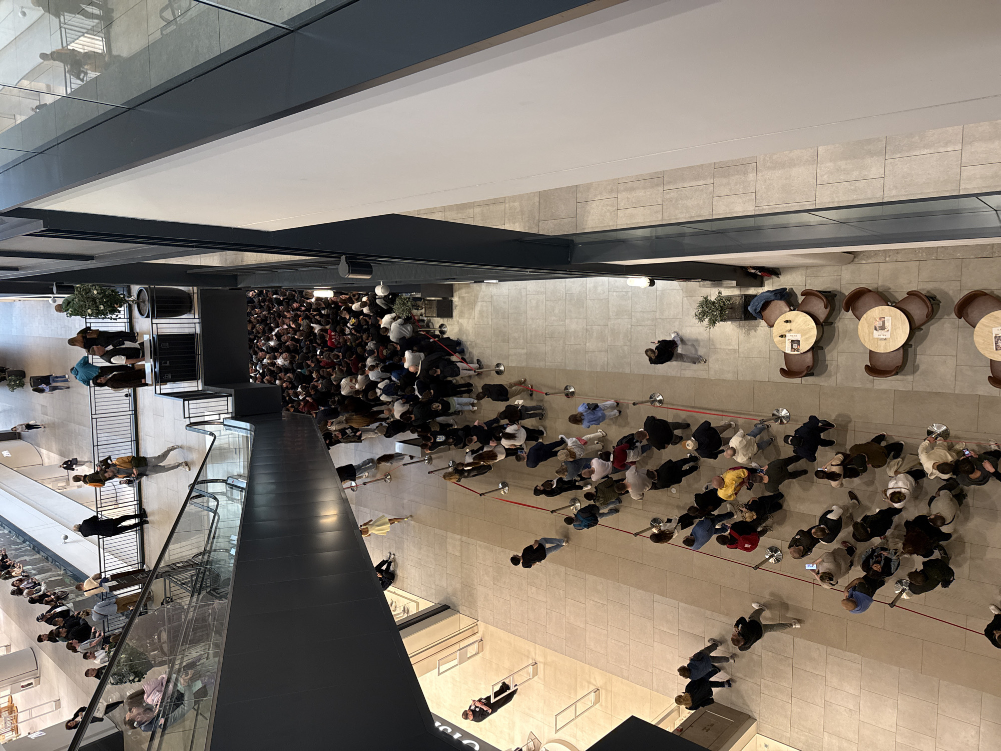 The image shows a large crowd of people queuing inside a building. The view is from above, looking down at the crowd. The location is a building in the city of Belgrade, Serbia. The queue stretches through a large open space with a tiled floor. There are tables and chairs to the right of the queue. The crowd is contained by red rope barriers. People are standing close together, waiting in line. Some people are looking at their phones. There is an escalator to the left of the image. A partial reflection of people above the crowd is visible in the glass barrier along the escalator.