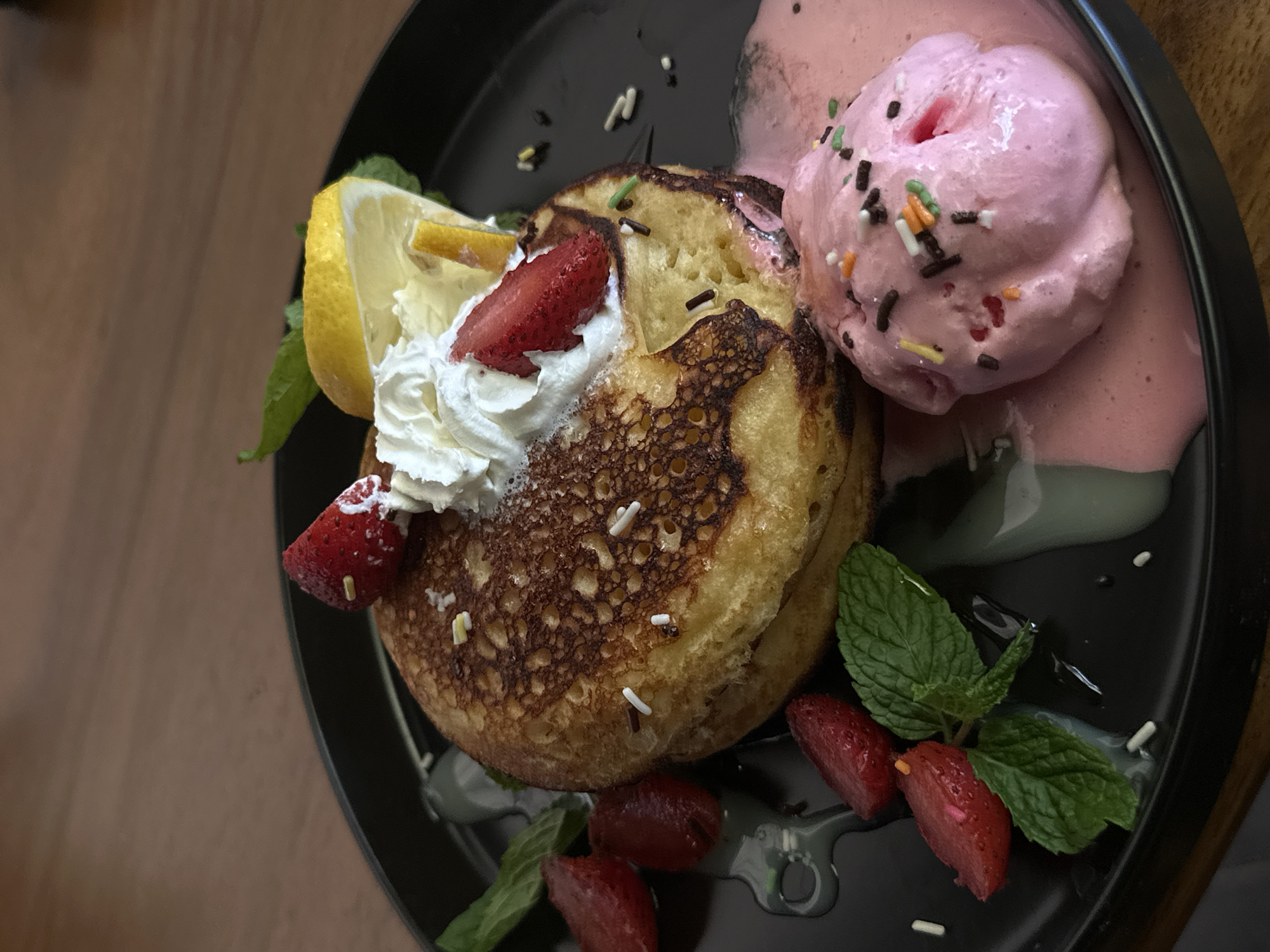 A close-up, eye-level shot captures a serving of pancakes on a black plate. The pancakes, stacked two high, are golden brown with a slightly crisp exterior and appear fluffy. They are topped with a dollop of whipped cream, a slice of strawberry, and a wedge of lemon. Sprinkles are scattered across the pancakes and the plate. To the side of the pancakes is a scoop of pink ice cream, also adorned with sprinkles. Sliced strawberries and fresh mint leaves are arranged around the pancakes, with a light syrup or sauce drizzled across the plate. The background shows a textured wooden surface. The lighting is dim, creating a warm, inviting atmosphere.