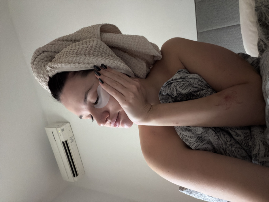 An adult female is depicted in a close-up, waist-up view within an indoor setting, consistent with a bedroom. Her head is wrapped in a light-colored, possibly beige, terry cloth towel. Two translucent, white hydrogel patches are applied beneath her closed eyes. Her left hand, featuring dark-painted fingernails, rests with fingers extended on her left cheek. Her bare right shoulder and upper chest are visible, partially covered by a grey and white patterned duvet or blanket. A reddish abrasion or mark is present on the inner aspect of her right bicep/tricep area. In the background, a white wall holds a wall-mounted air conditioning unit, which displays an illuminated green indicator light. A grey upholstered headboard and a white pillow are visible to the right. The lighting suggests ambient natural light illuminating the subject from the right. The exact location in Budva, Montenegro, cannot be determined from the visual information provided.