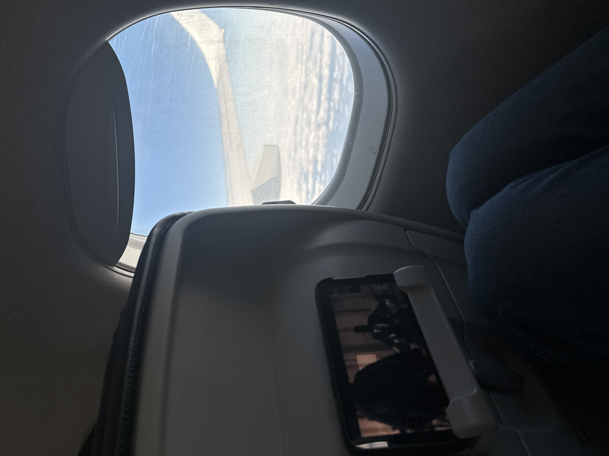 The image shows the interior of an airplane cabin. A passenger wearing blue jeans sits to the right. A seat-back tray table is folded down. A smartphone is positioned in the tray table's phone holder. Outside the airplane window, there is a view of the wing and the clouds. It is not possible to determine the location of Sant Josep de sa Talaia, Spain from the given image.