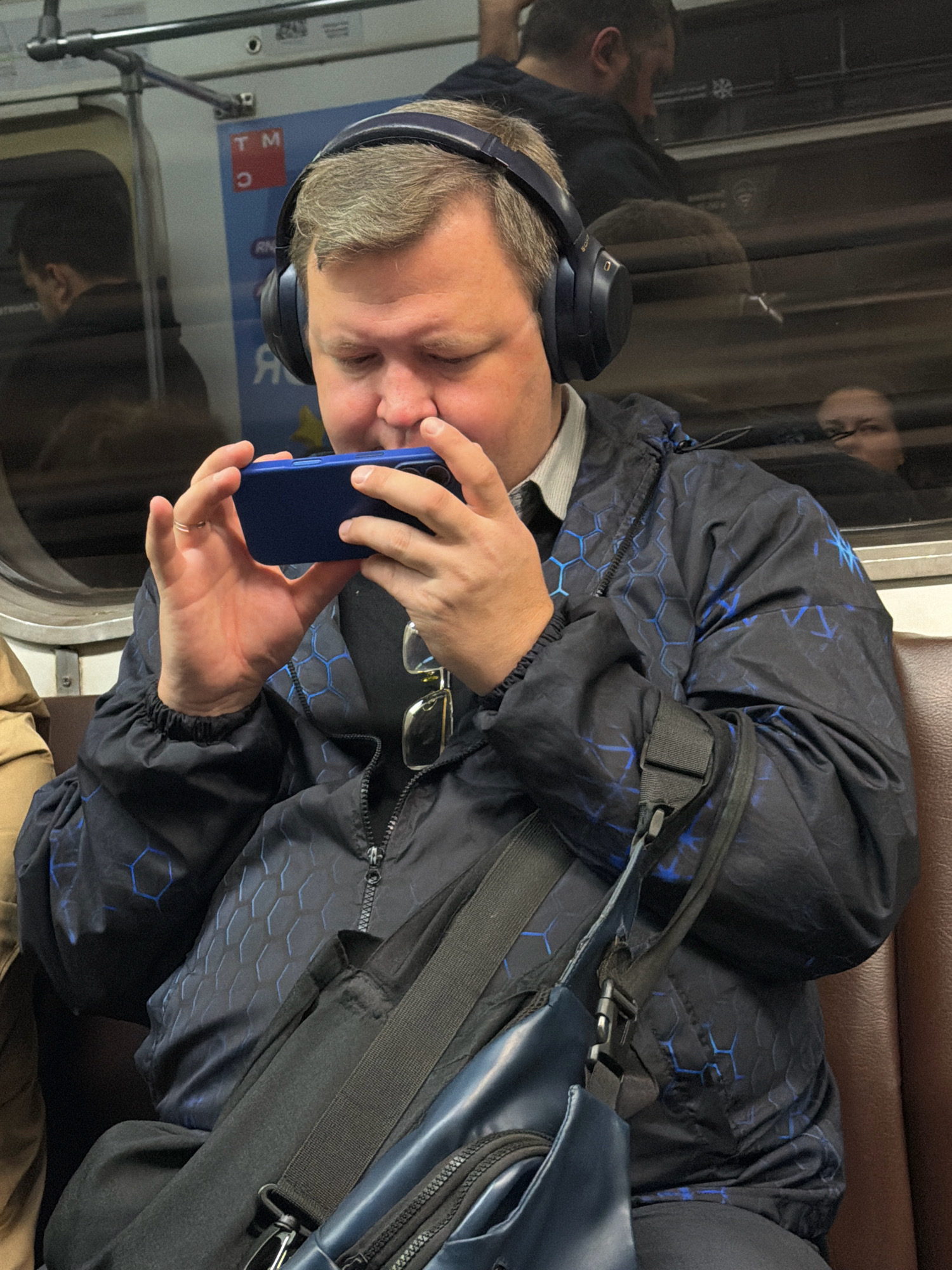 In Moscow, Russia, a man with headphones is seen sitting on a subway seat, engrossed in his smartphone. He is wearing a dark jacket with a blue hexagonal pattern and a light-colored collared shirt. He has a black bag slung across his chest. The subway car's interior is visible, including the window and seating. Reflections in the window show other passengers.