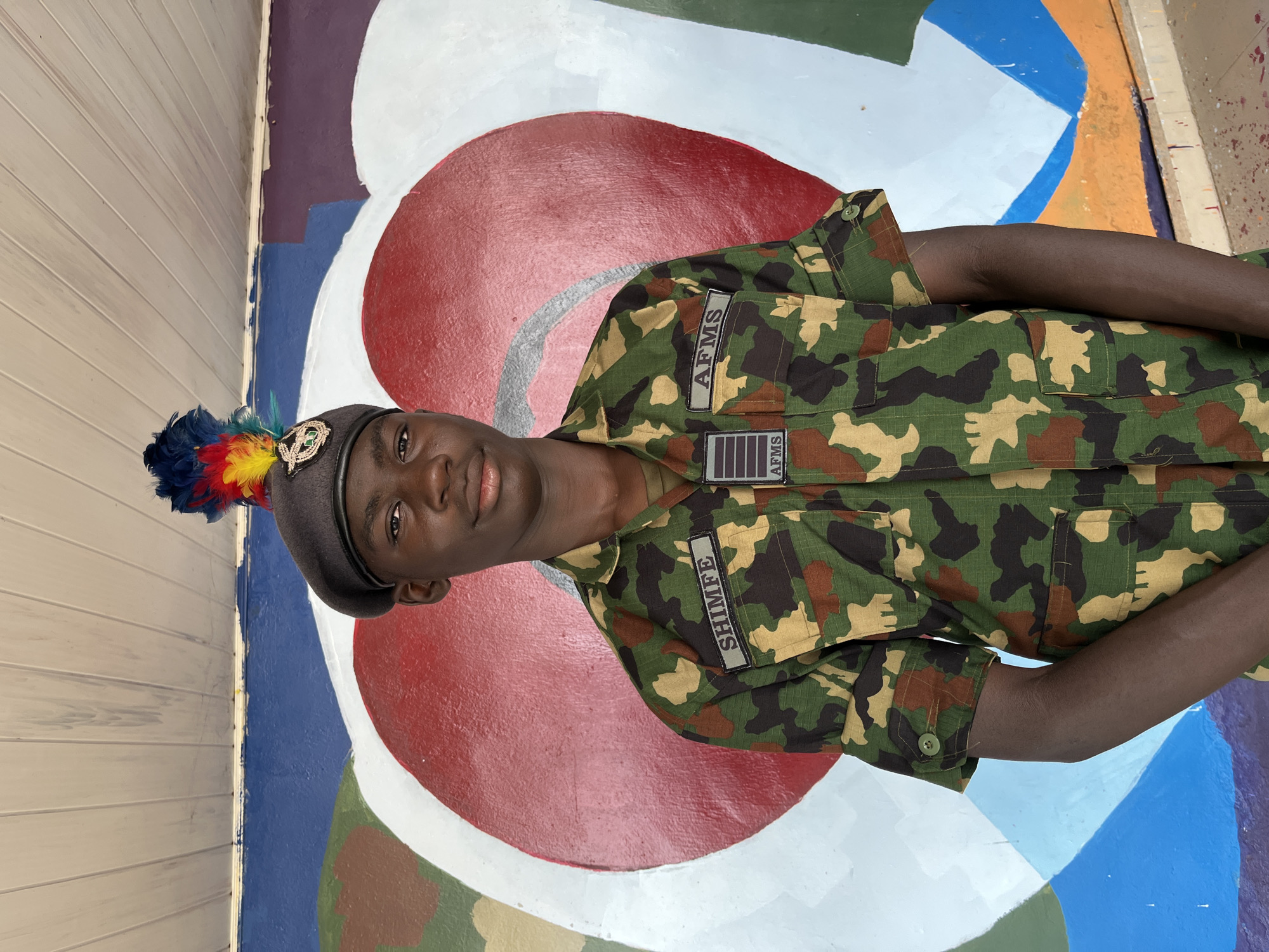 A young Black male, appearing to be in his late teens or early twenties, stands centered in the frame, facing the camera. He is wearing a camouflage military uniform with a dark beret adorned with a colorful feather plume. The uniform has patches on the chest, one reading "SHIMFE" and another "AFMS," with smaller insignia below the latter. He has a serious expression and is looking directly at the viewer. The background is a brightly painted mural featuring a large red heart with white and blue accents, set against a colorful abstract design on a wall. Above the mural, a white wooden ceiling is visible. The overall setting suggests an outdoor or semi-outdoor location, possibly in Bar Jirgi Summa, Nigeria, given the context. The lighting is even, indicating it is likely daytime.