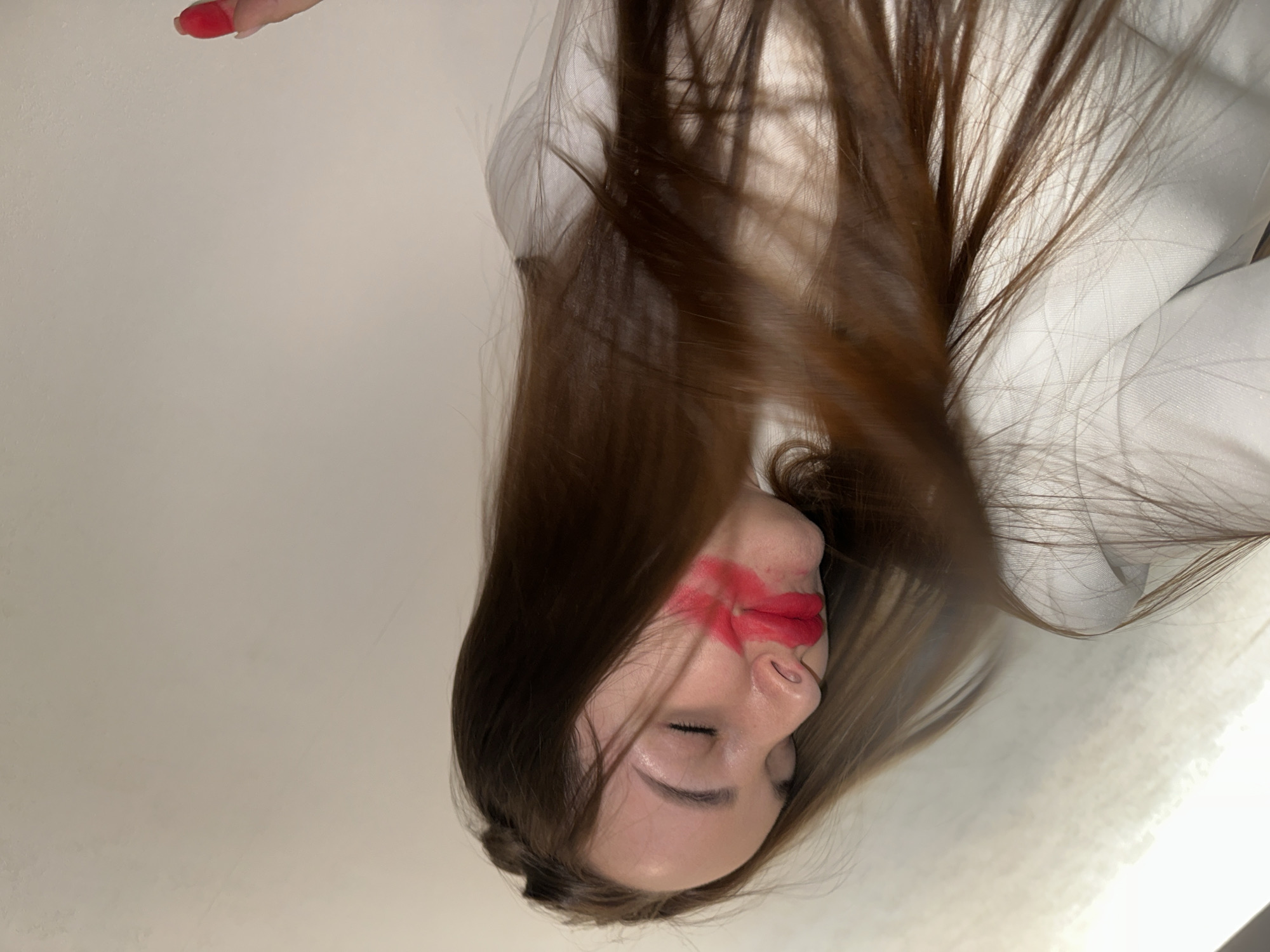 A woman with long brown hair lies with her face tilted upwards. She has red lipstick smeared around her mouth and cheek. Her eyes are closed. She appears to be wearing a white shirt or jacket. The background is a light gray wall or ceiling. The tip of a finger with red nail polish is visible in the upper left corner. There is no evidence to suggest the location is Chaikovsky, Russia.