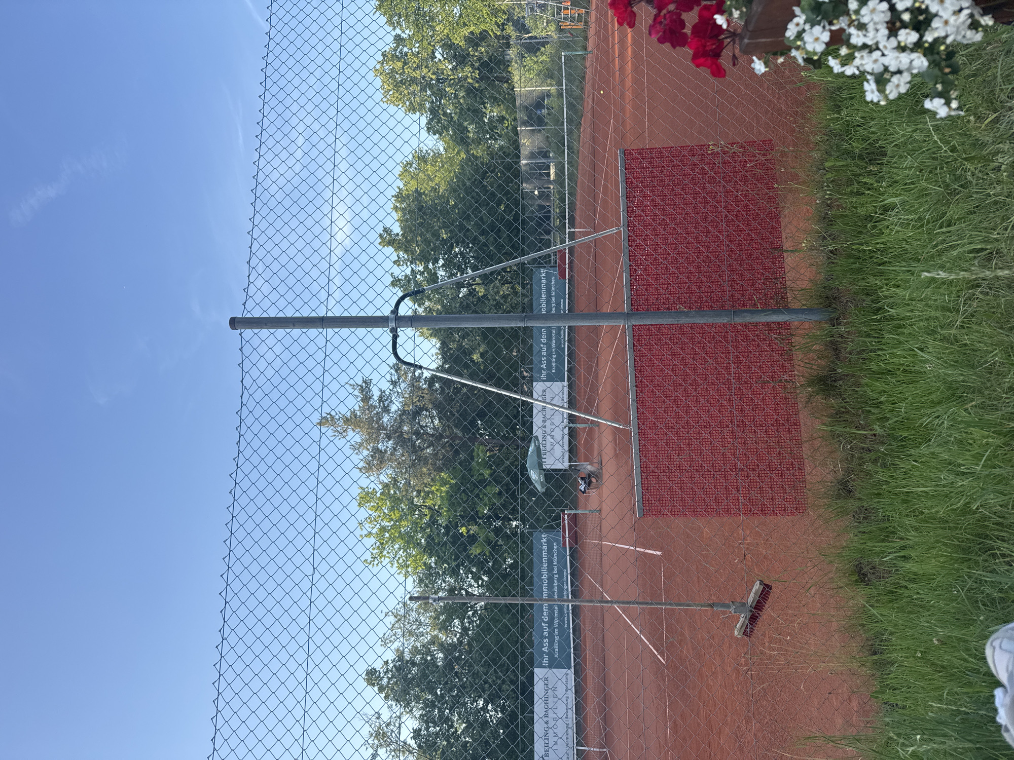 The image depicts an outdoor tennis court in Neubiberg, Germany, on a sunny day. The court is made of red clay, with white lines marking the boundaries. A green chain-link fence surrounds the court, with a metal pole supporting part of it. Behind the fence, a dense line of trees forms the background.
In the foreground, on the left side of the image, a broom is hanging from a pole attached to the fence. In the middle ground, a red windscreen is attached to the fence, partially obscuring the court. A green umbrella stands near the net on the right side of the court, and a person is sitting on a chair beneath it. A tennis ball is visible on the court in the right middle ground.
In the far right of the image, a wooden planter box is filled with red and white flowers. The sky above is a clear blue with a few wispy clouds. The overall impression is one of a well-maintained tennis facility on a pleasant day.