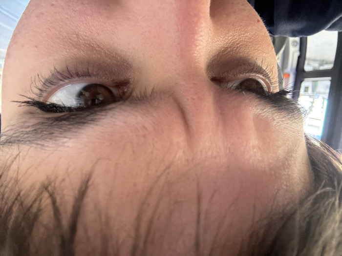 A close-up image depicts the upper portion of an individual's face, focused on the forehead, eyes, and upper nose. The subject has a furrowed brow, indicated by horizontal lines across the forehead and vertical lines between the dark, prominent eyebrows, suggesting a concentrated or stern expression. Both eyes are dark brown, with visible dark eyelashes that appear enhanced, and are directed towards the viewer's right. Dark hair is visible along the top of the frame. To the left of the subject's face, a dark vertical frame is present, likely a window pillar. Beyond this frame, an out-of-focus urban background is discernible under daylight, showing blurred light-colored architectural structures and indistinct shapes, consistent with a view from inside a vehicle, possibly public transportation. The specific location within the city of Sofia, Bulgaria, cannot be determined from the indistinct background elements.