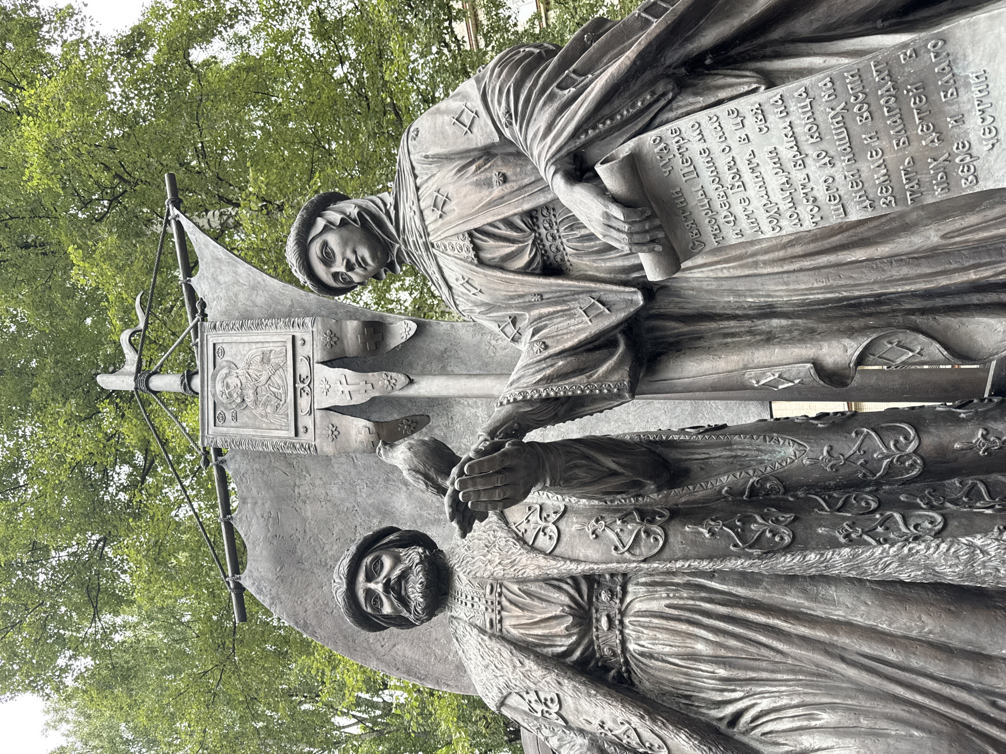 This is a low-angle, outdoor, medium shot of a bronze statue depicting two figures and a banner, set against a backdrop of lush green trees. On the left, a male figure with a beard, wearing a fur-trimmed robe and a headdress, holds a dove in his outstretched hand. To the right, a female figure, also adorned in elaborate clothing and a headdress, holds a scroll with Cyrillic text. Behind them, a large banner displays an icon at the top and ornamental designs. The statue appears to be located in Ladozhskoe Ozero, Russia. The lighting is overcast, creating soft shadows.