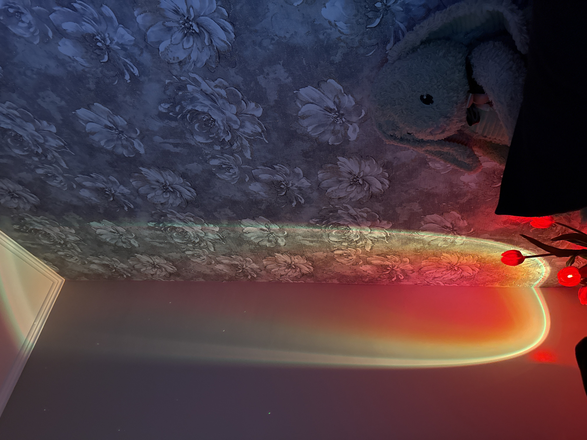 The image depicts a corner of a room with patterned wallpaper and a wall illuminated by a sunset lamp, casting a colorful gradient of light. In the foreground, a plush toy rabbit with a pink bow tie sits on a dark surface, partially obscured by shadows. Several red tulip-shaped lights are also visible, adding to the ambient lighting. The scene is devoid of any people and takes place in an indoor setting.