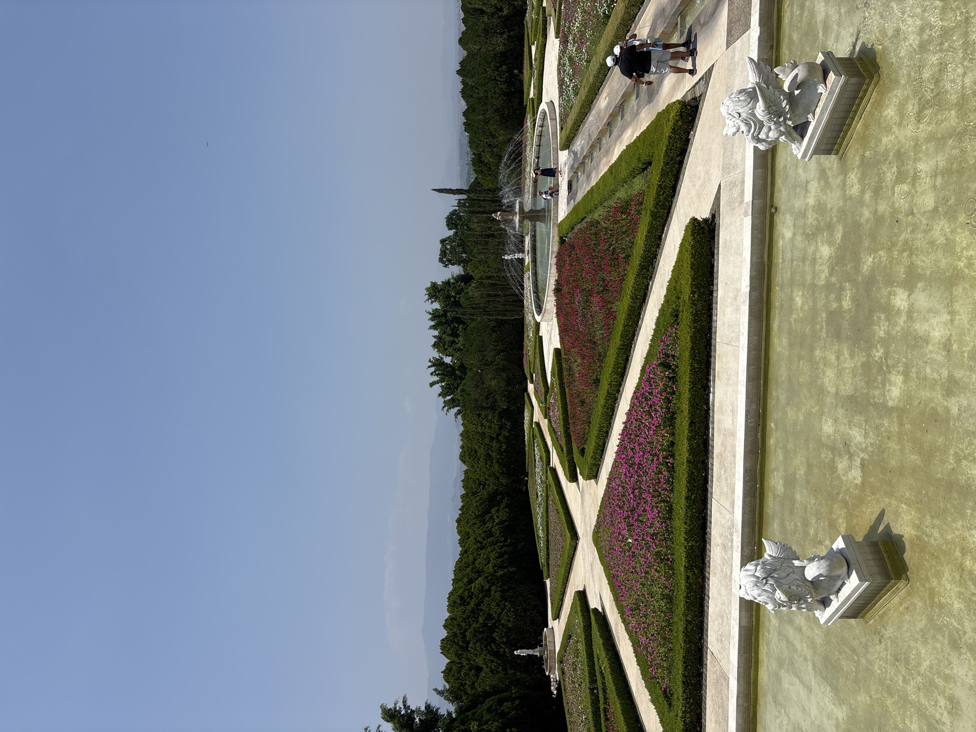 A slightly rotated, eye-level outdoor shot depicts a formal garden with geometric flowerbeds and pathways, likely situated in Tehuixtla, Mexico, given the prompt. A group of people is walking along one of the pathways in the background. Two stone statues, possibly depicting lions, are positioned along the edge of a rectangular pool of water. The water is tinted green. The garden is bordered by a dense line of trees. The sky is clear and blue.