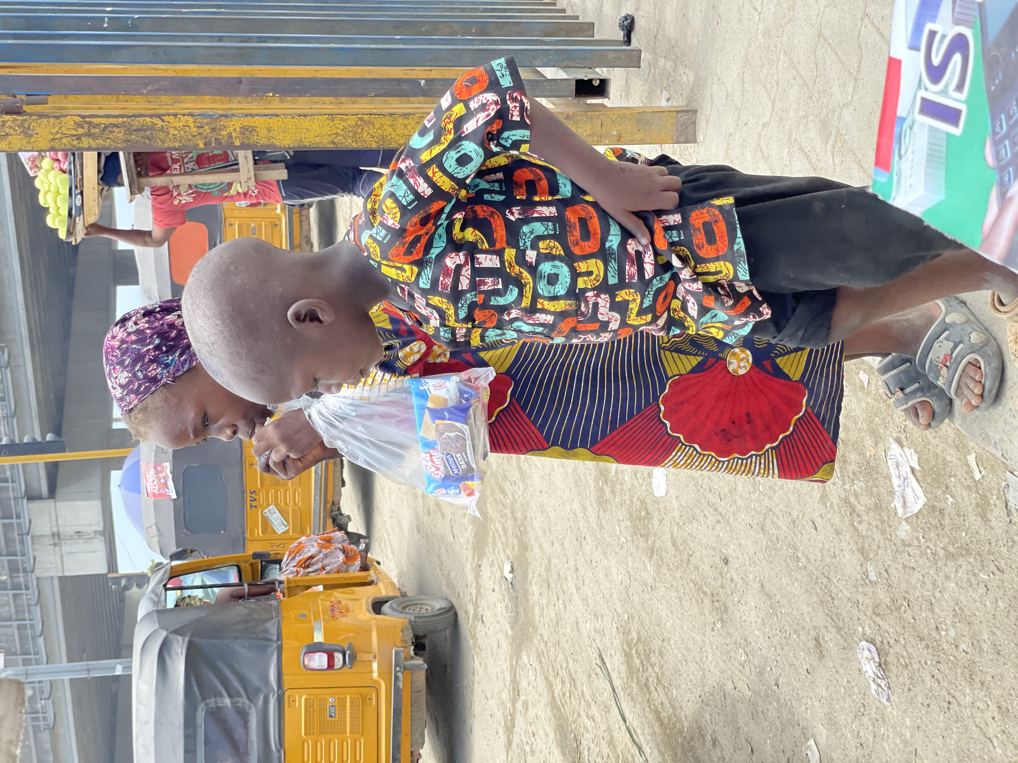 The image depicts a vibrant street or market scene in Lekki, Nigeria, under bright daylight. The primary subjects are a young child and an adult woman, both shown in profile.
The young child, with a bald head, is positioned on the left, looking downwards towards an item held by the woman. The child wears a distinctive black short-sleeved shirt covered in a colorful, repeating pattern of letters in various hues like orange, blue, yellow, and white. They also wear dark shorts and grey open-toed sandals.
Beside the child, an adult woman, likely an elder, wears a patterned purple headscarf and a long skirt made of a vibrant fabric featuring a large red scallop or shell-like motif on a dark blue background with golden stripes. She is holding a transparent plastic bag containing packaged cookies or biscuits. The packaging clearly shows text such as "MAYFAIR," "DAIRY," "MILK," and "BISCUITS."
The background reveals a bustling urban environment. Several yellow tricycles, known as Keke Napep, are visible, one bearing the "TVS" brand name. Another person in a red shirt stands behind a wooden stall, appearing to sell green fruits. Overhead, sturdy metal beams, painted yellow and blue, suggest an elevated structure or part of market infrastructure. The ground is a light-colored concrete or dirt surface, with scattered litter. A large sign or billboard, partially visible on the right, displays prominent white letters "IS" on a green background. The scene captures an everyday interaction amidst the activity of a Nigerian market setting.