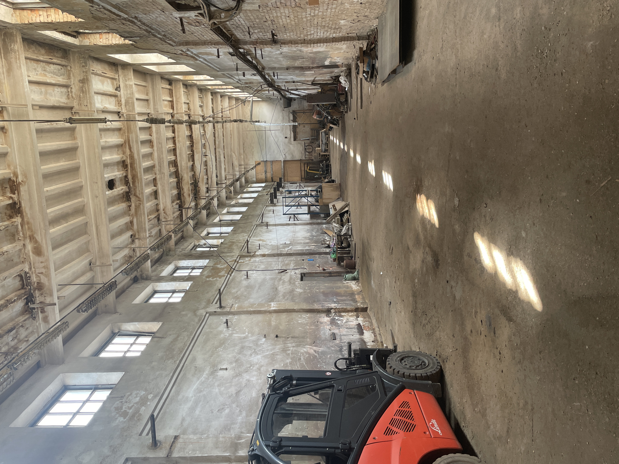 This media file depicts the interior of a large, industrial warehouse or factory in Saint Petersburg, Russia. The scene is oriented vertically, giving a sense of the height and depth of the space. The structure appears aged and possibly in disrepair, with exposed brickwork, weathered concrete walls, and a corrugated ceiling. Rows of tall windows line one side of the building, allowing natural light to stream in, casting elongated rectangular patterns on the dusty concrete floor. The other side of the warehouse is partially obscured by shadows and contains a jumble of stored materials, equipment, and what appear to be industrial machinery.
In the foreground, a bright orange and black forklift is prominently positioned. The forklift is stationary and appears to be in operational condition. The overall impression is one of a working or recently used industrial space, possibly undergoing renovation or simply in a state of organized clutter. There is no visible activity of people, suggesting the image was captured during a quiet period or when the space was empty. The lighting suggests it could be daytime, with sunlight filtering through the windows. The air quality appears dusty, consistent with an industrial or storage environment. No text is readily visible on any of the objects within the frame.