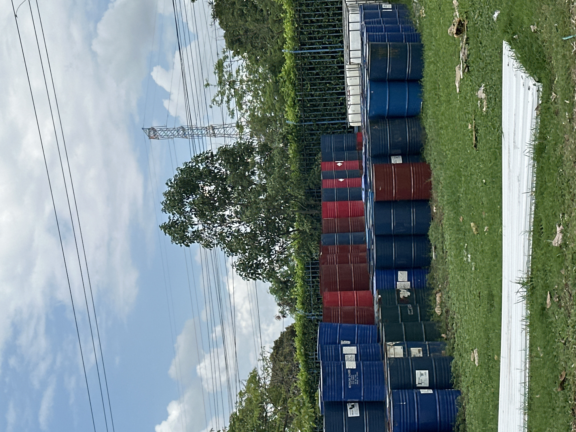 The image shows an outdoor scene with stacks of metal barrels in Juan Mina, Colombia. The barrels are predominantly blue, with some red and green ones interspersed. They are arranged in rows, with some stacked two high. In the background, there is a fence, lush green trees, and a transmission tower against a partly cloudy sky. The ground is covered with grass and some scattered debris. There are no people or animals visible in the image. The overall impression is of an industrial or storage area.