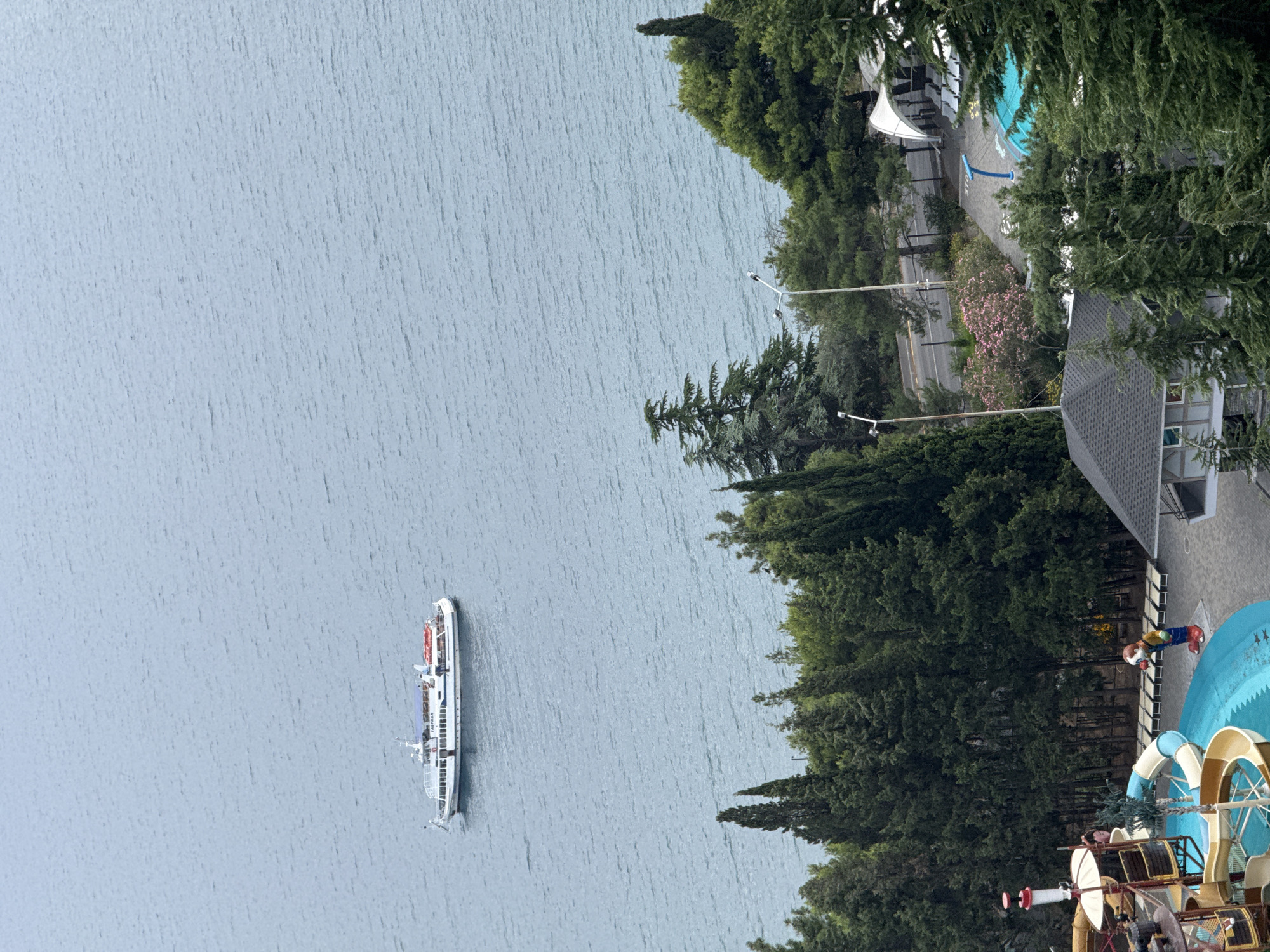 The image depicts a scenic view of the sea and a coastal area, likely from an elevated vantage point, possibly within or overlooking the city of Yalta, Ukraine. In the distance, a white passenger boat with a blue awning is navigating the calm, light blue sea. Closer to the shore, a dense line of dark green cypress and other coniferous trees forms a natural barrier. Beyond the trees, a road is visible with a few parked cars. There are also indications of recreational facilities, including lounge chairs and a section of a turquoise swimming pool with a blue water slide. A figure dressed in colorful clothing, possibly a park employee or a statue, stands near the pool area. In the foreground, a water park attraction with slides and structures is partially visible, along with a small building. The overall atmosphere is tranquil, with a blend of natural landscape and man-made amenities.