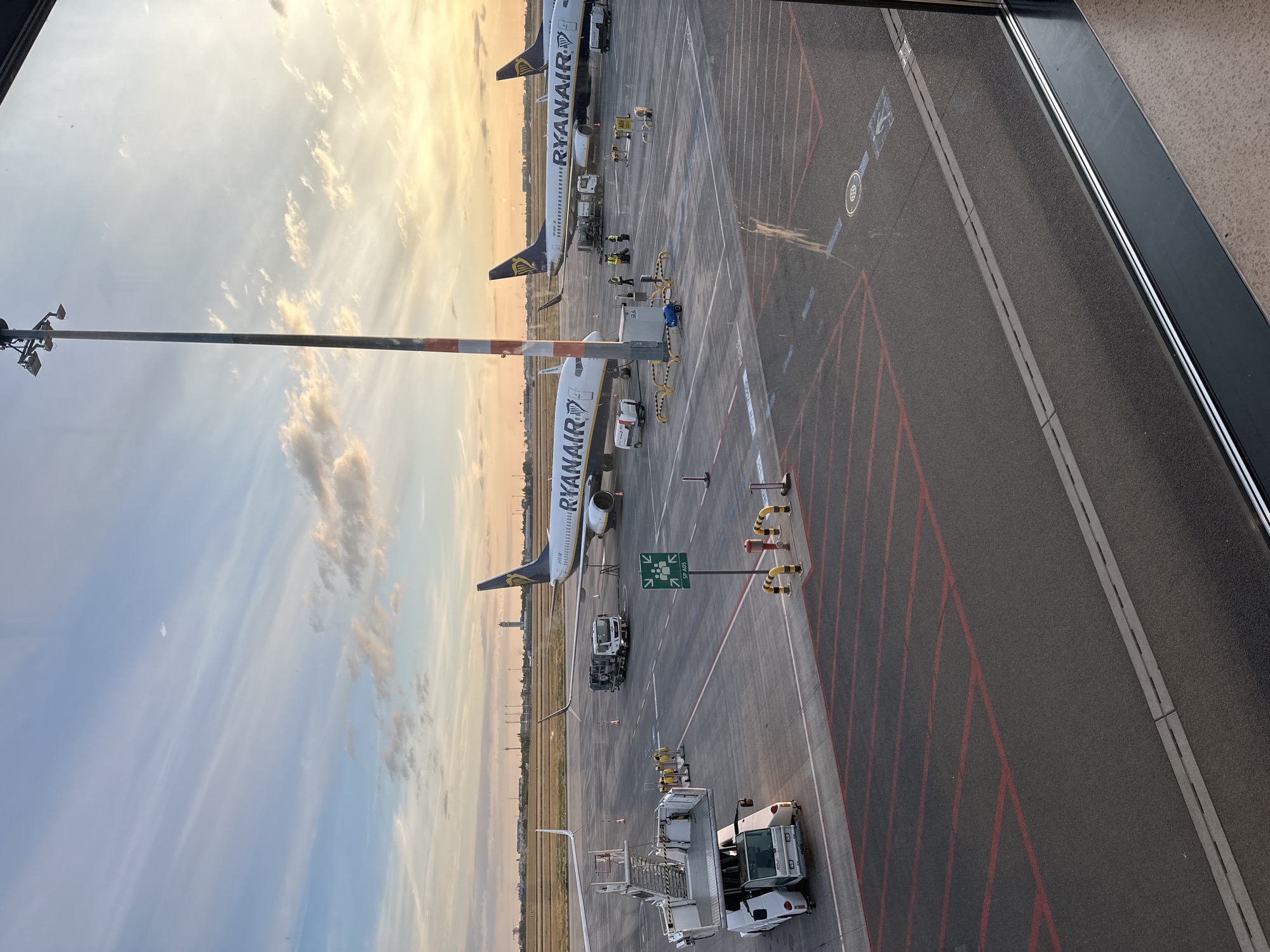 In the city of Schönefeld, Germany, at an airport, two Ryanair airplanes are parked on the tarmac. The sky is filled with wispy clouds, illuminated by the golden light of a sunset. A tall lamppost with lights stands prominently on the right side of the image. In the foreground, there is a large white vehicle, possibly a baggage loader, parked on the asphalt. Red lines mark parking spots on the tarmac. Several airport personnel in yellow vests are visible near the airplanes, attending to their duties.