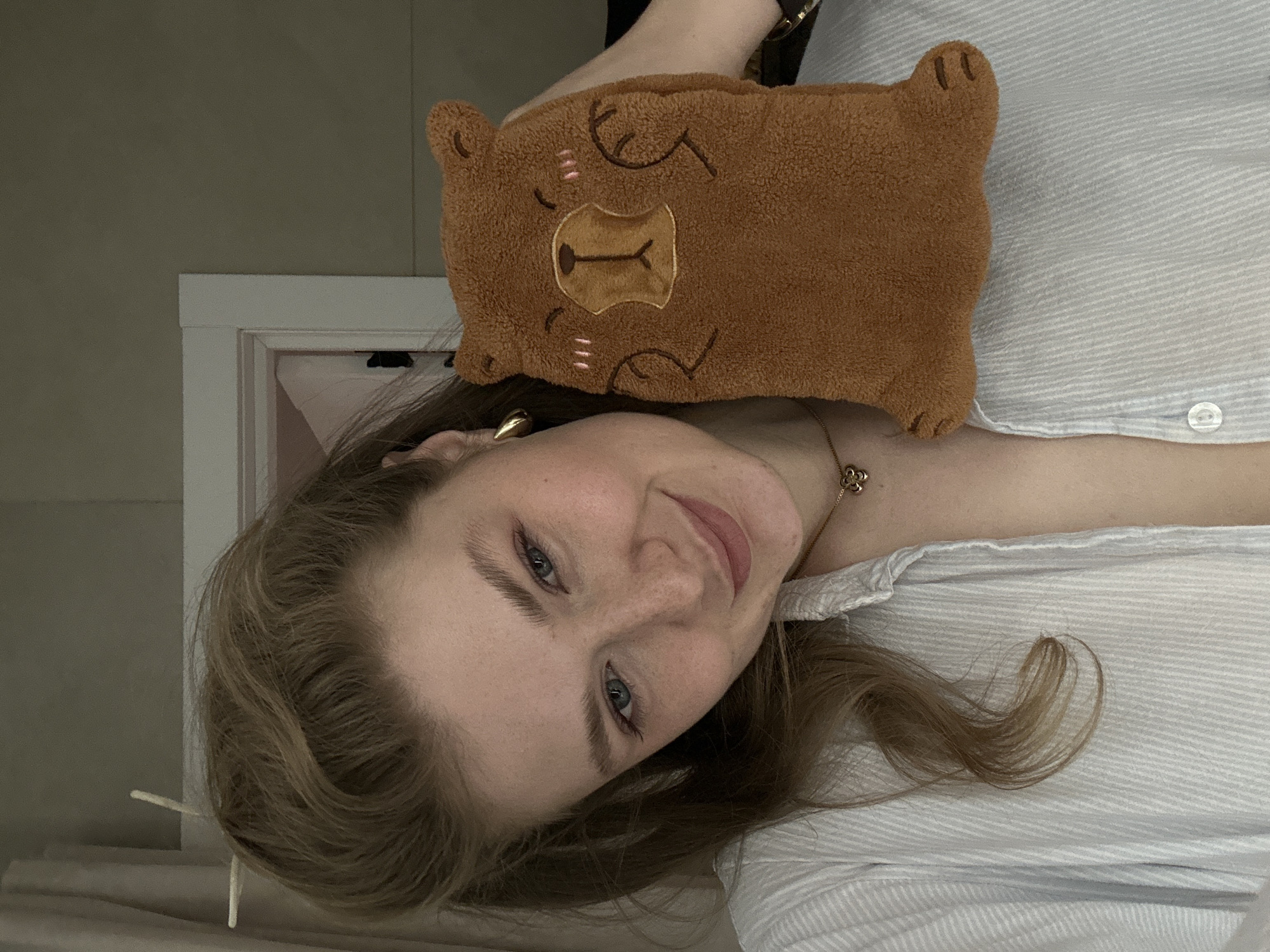 A young woman with fair skin, light brown hair, and blue eyes is holding a brown, square-shaped, plush bear-like object to her head. She is wearing a light-colored, button-down shirt. A gold necklace is visible around her neck. Part of a white doorway is visible in the background. The location is unknown and there is no evidence to connect it to Kazan, Russia.