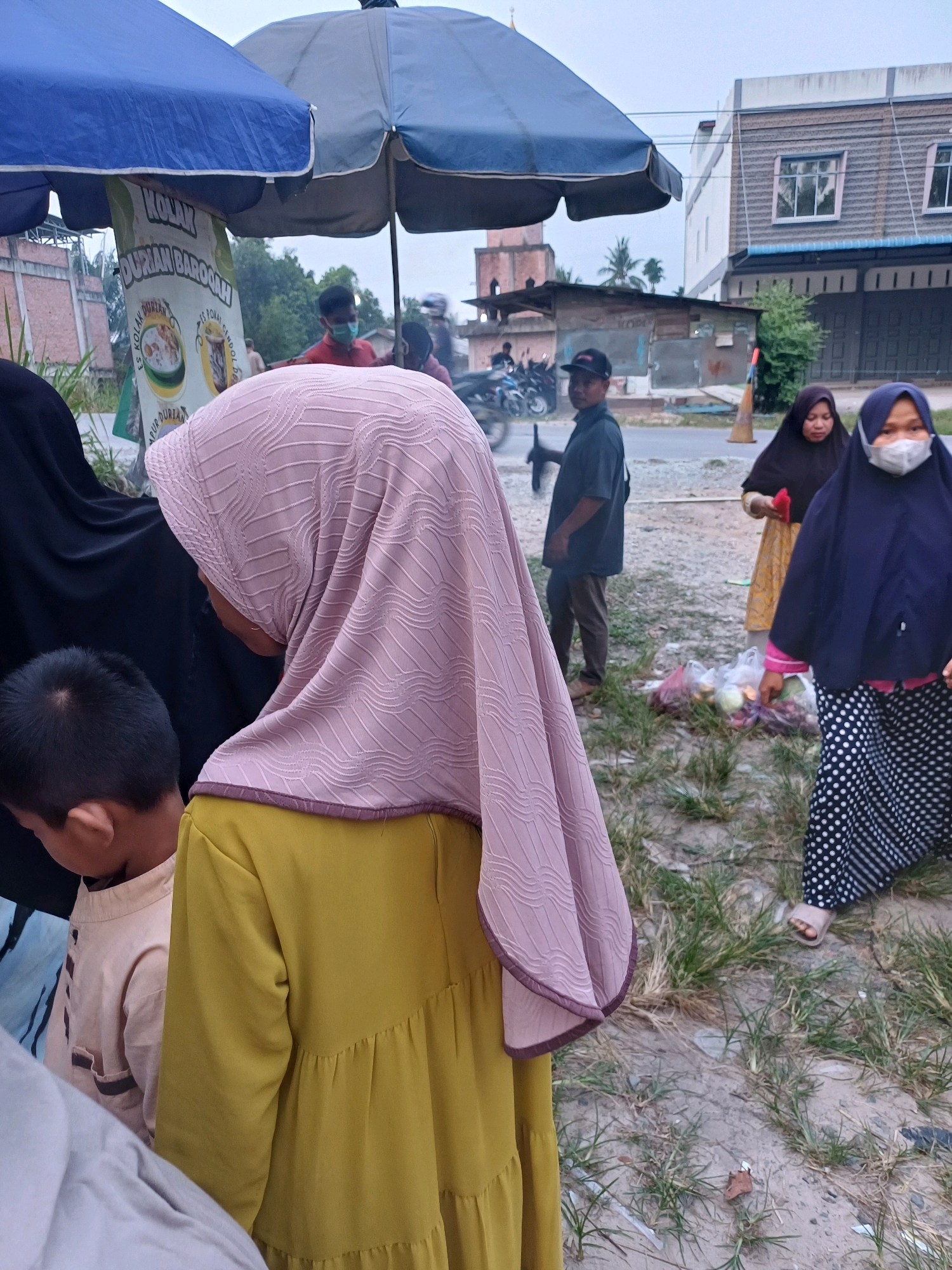 This outdoor scene depicts a roadside stall in Dumai City, Indonesia, on an overcast day. In the foreground, a child wearing a light brown shirt is partially visible, looking down. To the right, a woman in a yellow dress and a light pink hijab is also seen from the side. Further to the right, another woman is walking away, wearing a dark blue hijab and a black and white polka dot skirt, with a medical mask covering her face. Behind her, a woman in a dark hijab and a patterned yellow and orange dress stands near some plastic bags containing produce.
A man in a dark green shirt and a black baseball cap stands in the middle ground, facing away from the camera. Behind him, a stall with blue and grey umbrellas is set up, displaying a banner with text that includes "KOLAK DURIAN BAROQAH". Several motorcycles are parked near the stall. In the background, there are buildings with windows and a mosque-like structure with a minaret. The ground is a mix of dirt, gravel, and patches of grass. The overall impression is of a modest commercial setting in a moderately populated urban area.