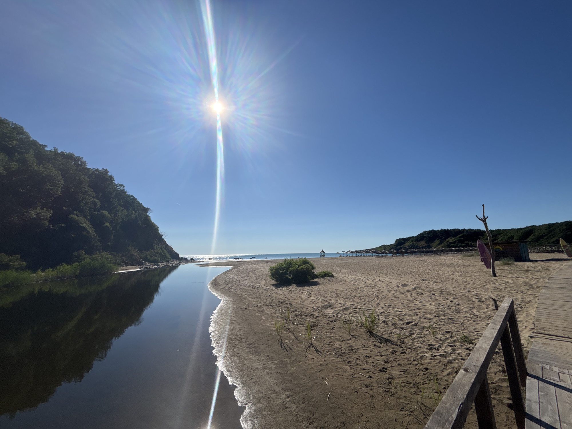 A bright, sunny day at a beach in Rezovo, Bulgaria. A calm, dark body of water, likely a river or lagoon, flows from the left side of the image towards the sea. The water reflects the lush green trees and vegetation covering the steep hill that borders it on the left. The beach is sandy and extends to the right, meeting the gentle waves of the sea. In the distance, several beach umbrellas and a small wooden structure are visible, suggesting a developed beach area. A wooden boardwalk leads from the lower right corner of the frame towards the beach. The sun is high in the sky, casting bright light and lens flares across the scene. There are no people visible in the image.