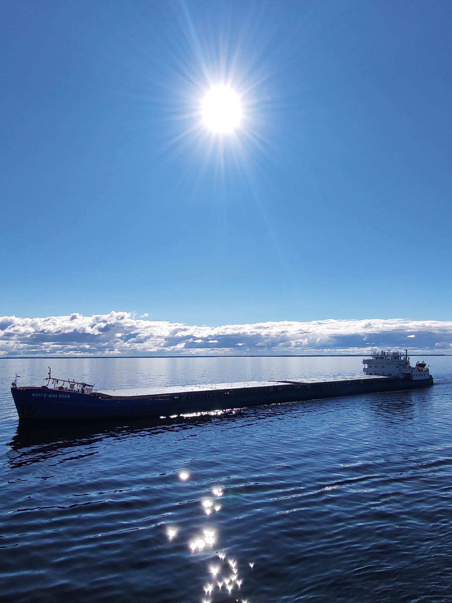 A medium shot captures a ship sailing on a body of water beneath a bright, sunlit sky. The ship, identified as "ВОЛГО-ДОН 5068," is a large, flat-topped vessel moving from left to right in the frame. The sky is mostly clear blue with a few scattered clouds near the horizon. Sunlight glints off the water's surface, creating bright highlights. The scene appears to be a natural, outdoor environment, possibly a river or large lake near Novaya Svobodka, Russia. There is no evident human activity beyond the vessel's operation.