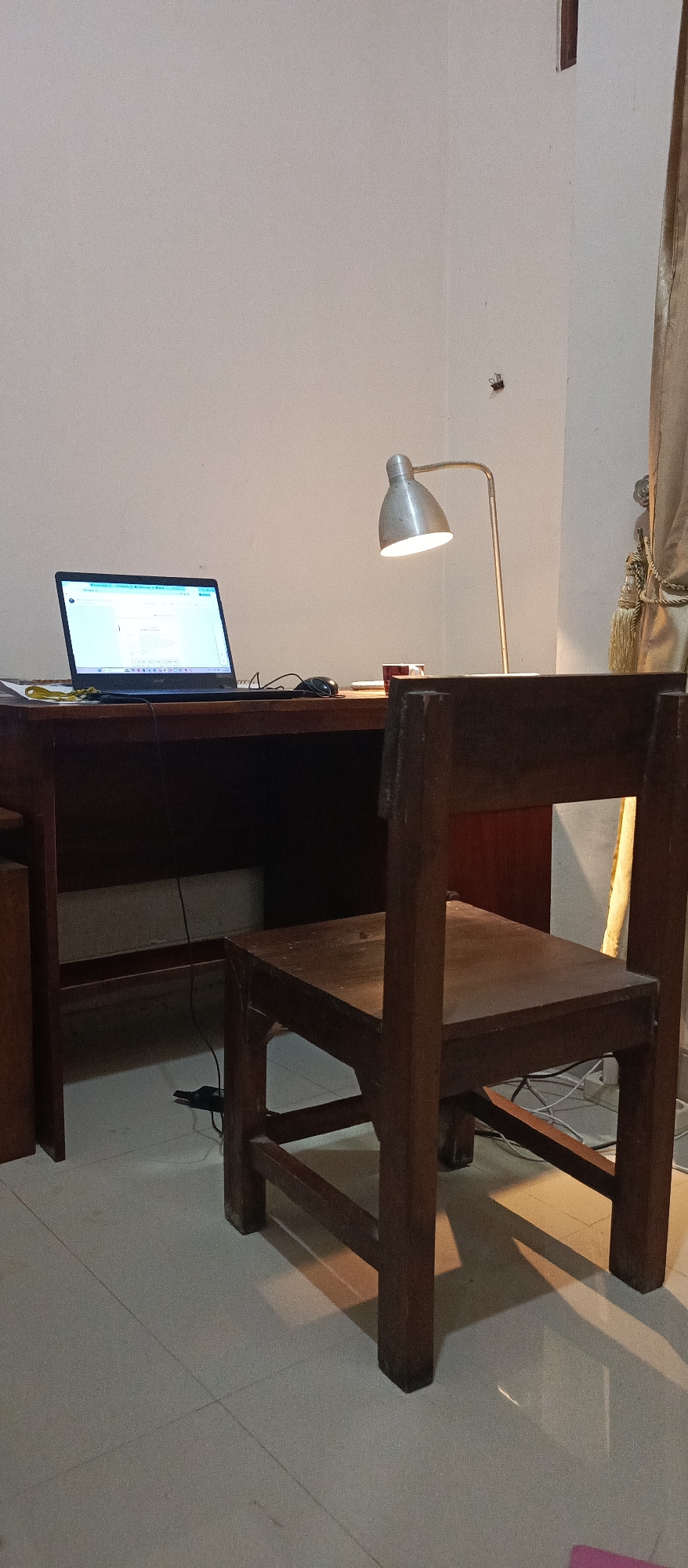 The image shows a wooden desk with a laptop and a desk lamp on it. A wooden chair is placed in front of the desk. The scene appears to be an indoor study or workspace, possibly in Jatisari, Indonesia, given the context. The desk lamp is switched on and casting a warm light. A computer mouse and a small mug are also visible on the desk. The floor is tiled. The overall impression is that of a simple, functional workspace.