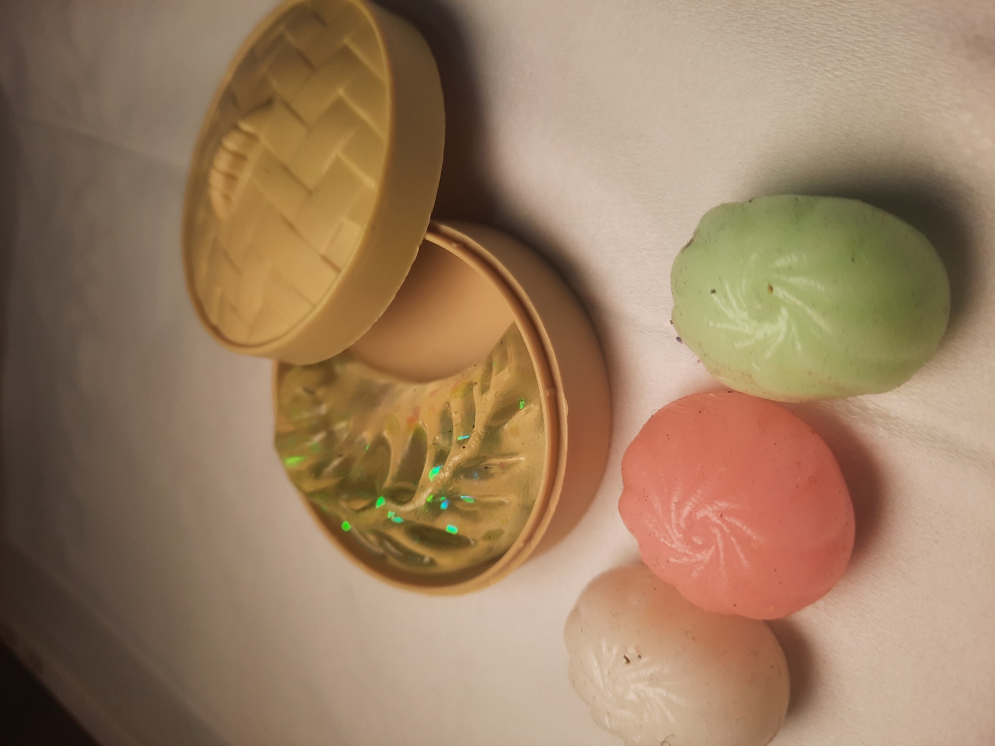 A still life photograph shows three molded objects resembling dumplings or buns, arranged on a textured white surface. To the right of these objects is a light brown, round container with a lid, designed to resemble a bamboo steamer. Inside the container is a yellowish-green, wavy object, possibly representing food, with several small, bright green and blue specks scattered on its surface. The lighting is dim and directed from the top, creating shadows and highlighting the textures of the objects. The background is out of focus and appears to be a neutral, muted color. The image does not depict any specific location in North Jakarta, Indonesia, or any identifiable people or actions.