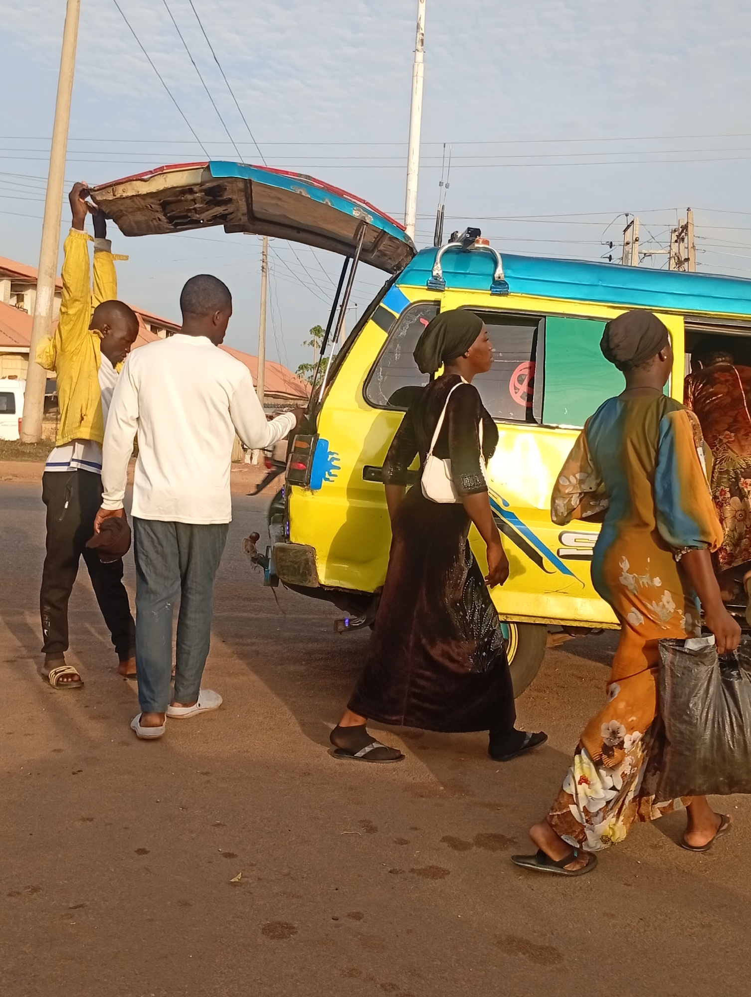 In Barden Yana, Nigeria, a yellow and blue minibus taxi is stopped on a paved street. The vehicle's rear door is open and being held aloft by a man in a yellow jacket. Another man in a white shirt stands nearby. Two women in dark head coverings are walking past the vehicle, with one carrying a white handbag and the other holding a black plastic bag. The background includes buildings and utility poles.