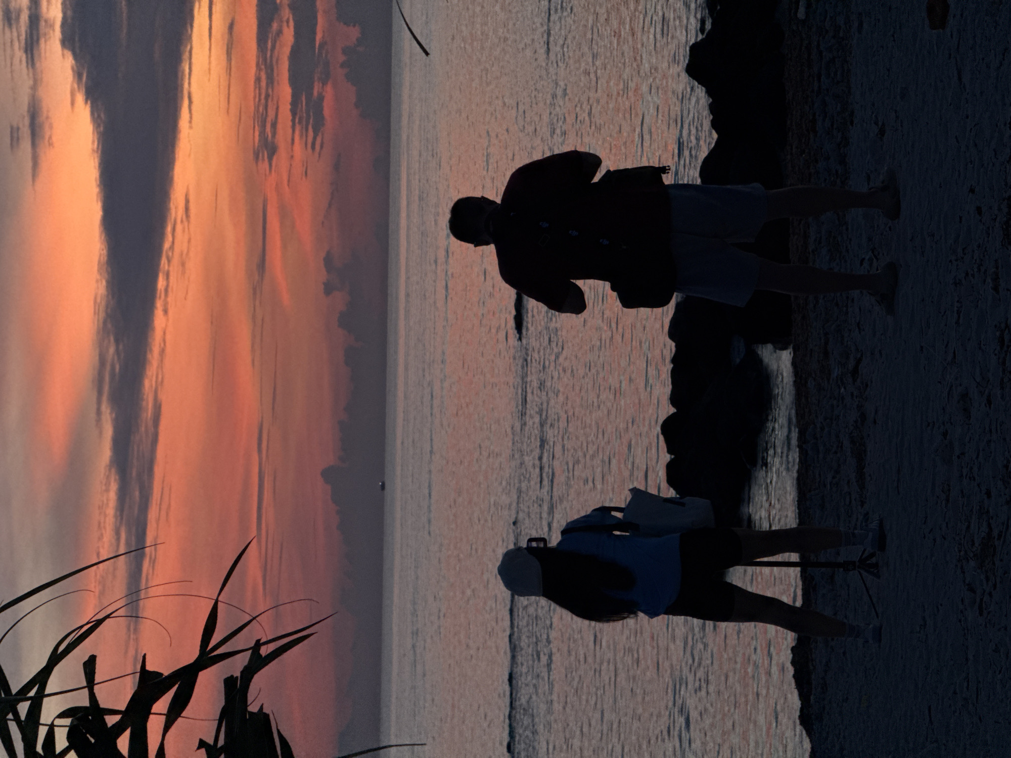 Two people stand on a sandy beach, silhouetted against a vibrant sunset over the ocean. A woman, facing away from the camera, wears a cap and a light-colored shirt with dark shorts, and is holding what appears to be a phone or camera. A man, also facing away, wears a dark t-shirt and light shorts, with a camera bag slung over his shoulder. The ocean is calm, with gentle waves lapping the shore. In the distance, a small boat is visible on the water. The sky is a dramatic display of orange, pink, and purple clouds, characteristic of a tropical sunset. The scene likely takes place in the coastal area of Lý Sơn Special Administrative Region, Vietnam, known for its beautiful beaches and sunsets.