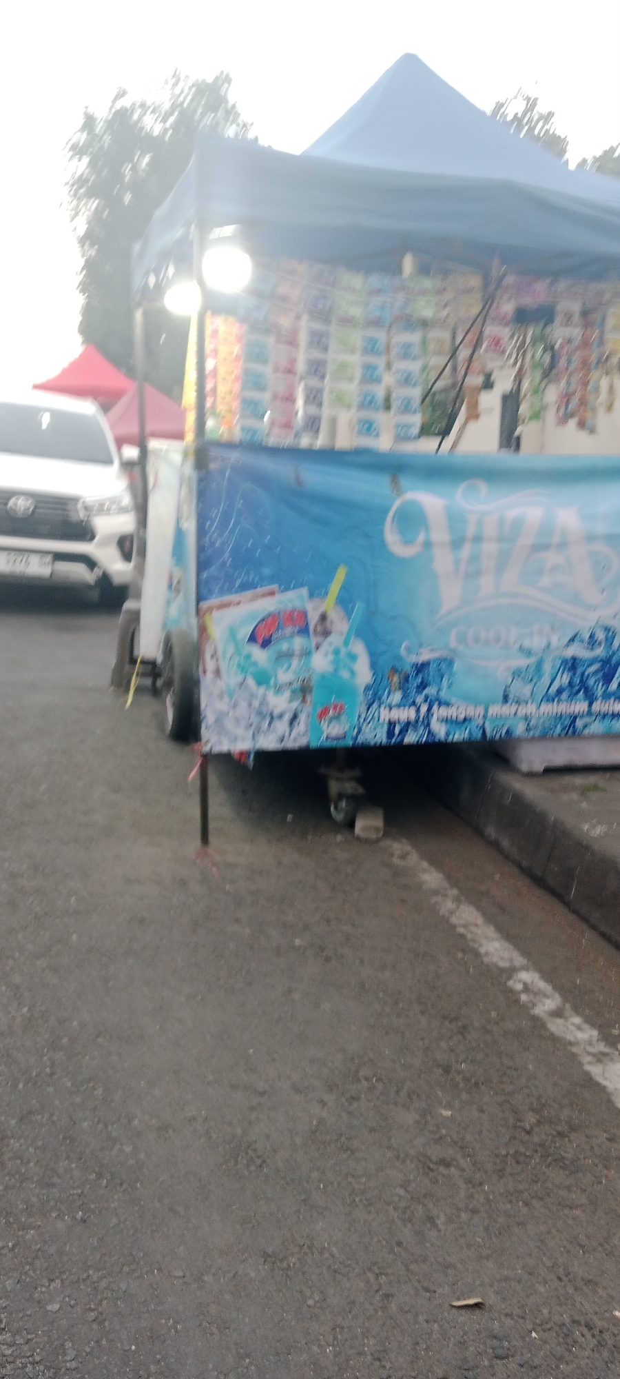 A white Toyota Innova with license plate B 2776 GBA is parked on the left side of the image, facing right. Behind it, a red tent structure is visible. A food cart with a blue tarp is parked on the right side of the image, facing forward. The tarp features a graphic with the word "VIZA" in large white lettering and images of blue drinks with straws. The cart is supported by a black pole on the left and has wheels on the right. The ground is paved and appears to be a road with a white line marking. The background shows trees and a bright sky. The scene is likely an outdoor market or roadside stall in Ngemplak, Indonesia.