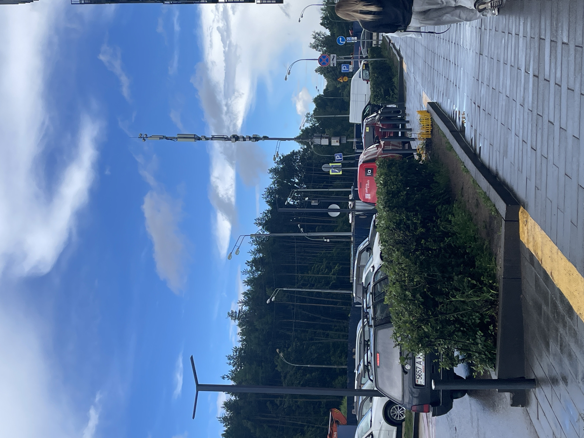 A street scene in Moscow, Russia, on an overcast day. A cellular tower stands tall against a blue sky with scattered clouds, surrounded by trees. The wet pavement suggests recent rain, with puddles reflecting the sky. Several cars are parked along the street, and a woman with blonde hair is walking away from the camera on the right side of the image. The ground is paved with gray tiles.