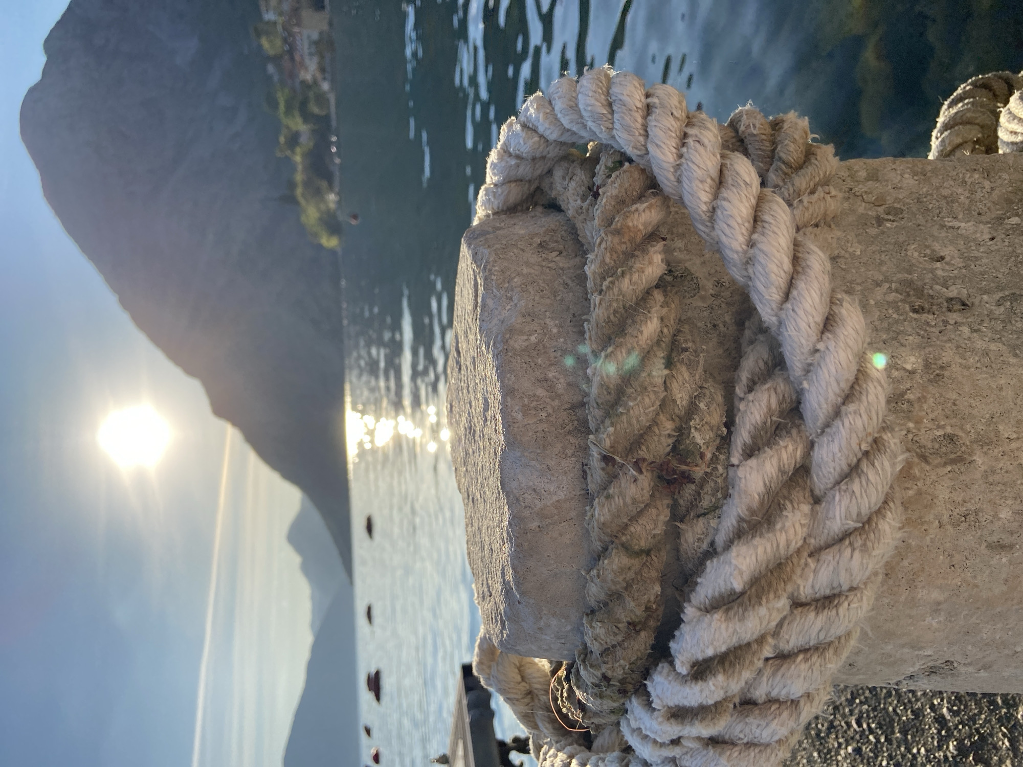 A rope is coiled around a gray concrete bollard at the edge of the Bay of Kotor in Donji Orahovac, Montenegro. The water is visible behind the bollard, reflecting sunlight. Mountains are silhouetted in the background, with the sun partially obscured.