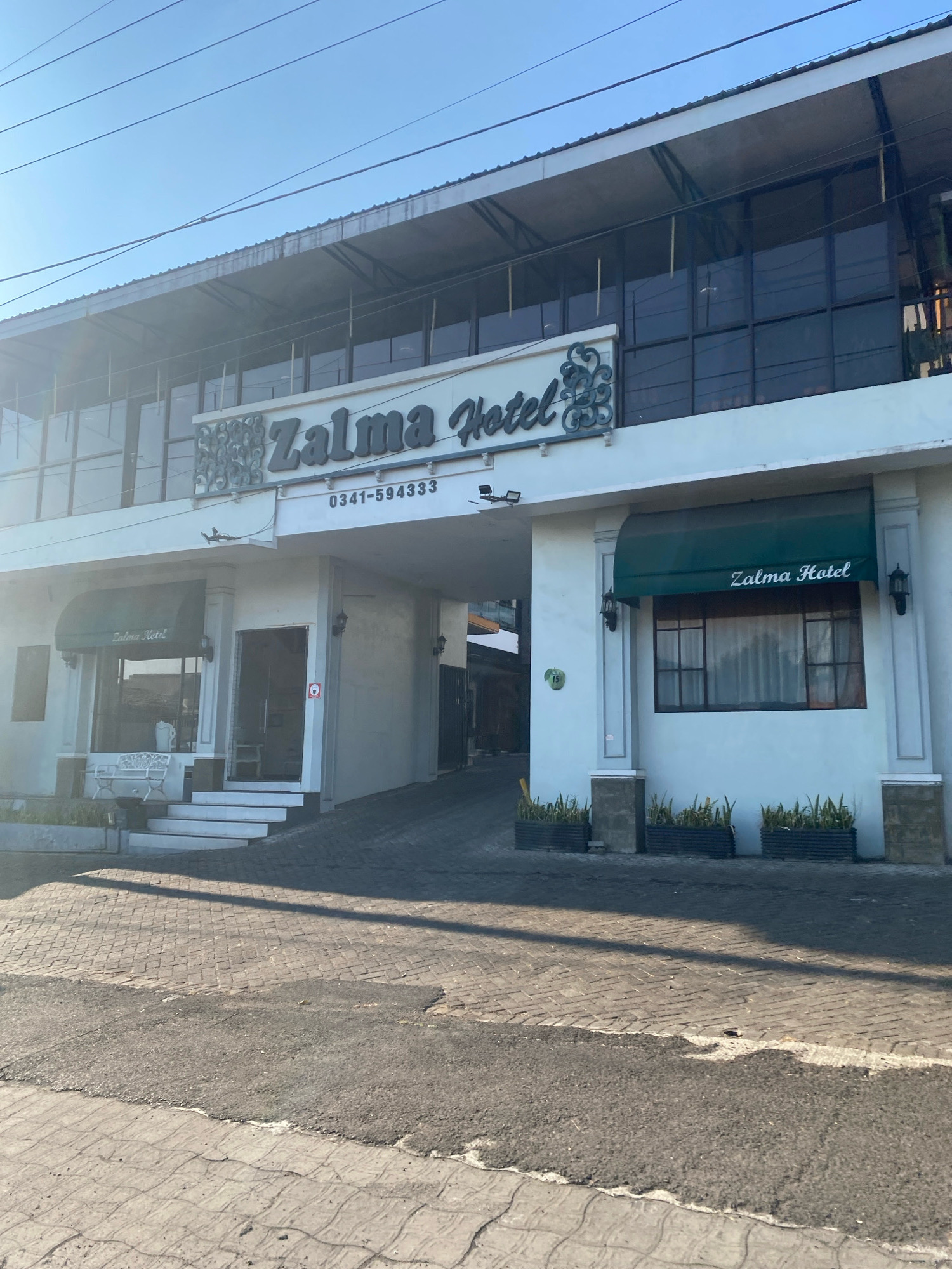 The image shows the exterior of the Zalma Hotel, a two-story building with a white facade and a dark-tiled roof. The hotel entrance is an archway leading into the building, with a sign above that reads "Zalma Hotel" in large, stylized letters. Below the sign, a phone number is visible: "0341-594333".
On the left side of the entrance, there is an awning with "Zalma Hotel" written on it, and a white bench sits outside. A set of white stairs leads up to the entrance. On the right side of the entrance, there is another awning with "Zalma Hotel" on it, and a window with curtains. Several potted plants are placed in front of the building on this side.
The building is situated on a street paved with cobblestones and asphalt. The sky is clear and blue. The overall scene appears to be in a city, likely in a warm climate, given the clear sky and outdoor seating.