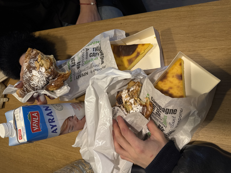 The image displays a light brown wooden tabletop laden with food and beverages, suggesting an indoor setting in Asnières-sur-Seine, France. Two individuals are present, evidenced by visible hands and lower arms. On the left, a hand with a dark sleeve is reaching into a white plastic bag containing a partially consumed almond croissant, dusted with powdered sugar and wrapped in white paper printed with French bakery terms such as "BONJOUR" and "LEVAI N à emporter". Adjacent is a partially visible clear, ribbed plastic bottle. Centrally located is a full carton of "Yayla Ayran" yogurt drink with a white screw cap, featuring an image of a person drinking. To the right, a second hand with light-colored manicured fingernails and a dark, possibly furry, sleeve holds another partially eaten almond croissant, similarly powdered. A red string bracelet is visible on this person's wrist. Also visible are two triangular slices of flan pâtissier (custard tart) in white cardboard containers, one fully exposed, the other partially within a white plastic bag, both displaying a caramelized top surface. The specific establishment within Asnières-sur-Seine is not identifiable from the image content.
