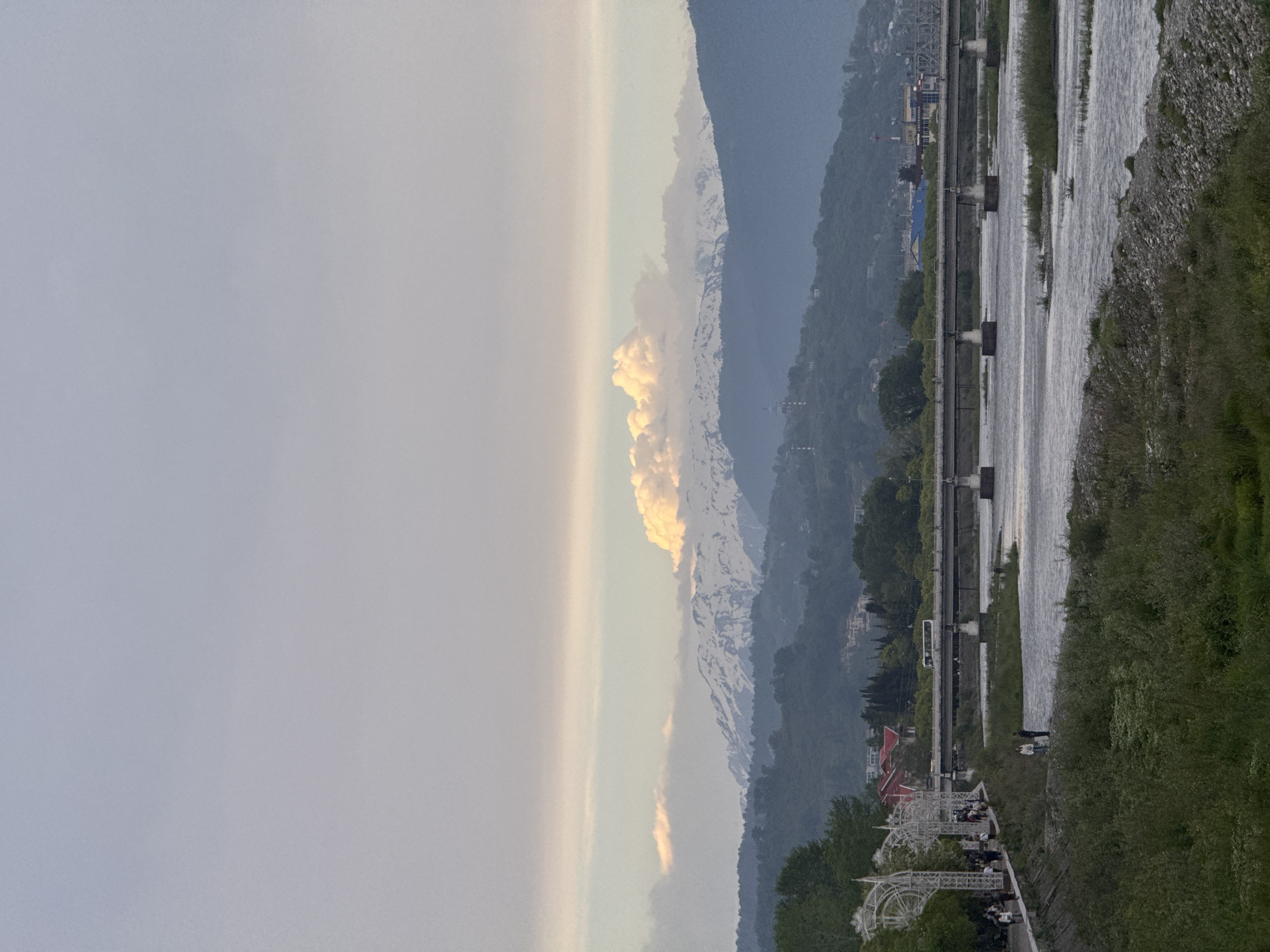 The image shows a landscape with mountains in the background and a river in the foreground. The sky is cloudy with patches of light. Some snow is on the mountains. A walking path with people on it is on the left of the river. A bridge or road is parallel to the river. A forensic analysis suggests the image may depict the Mzymta River and surrounding area in Sochi, Russia, which features mountainous terrain near the city. Some people are walking.