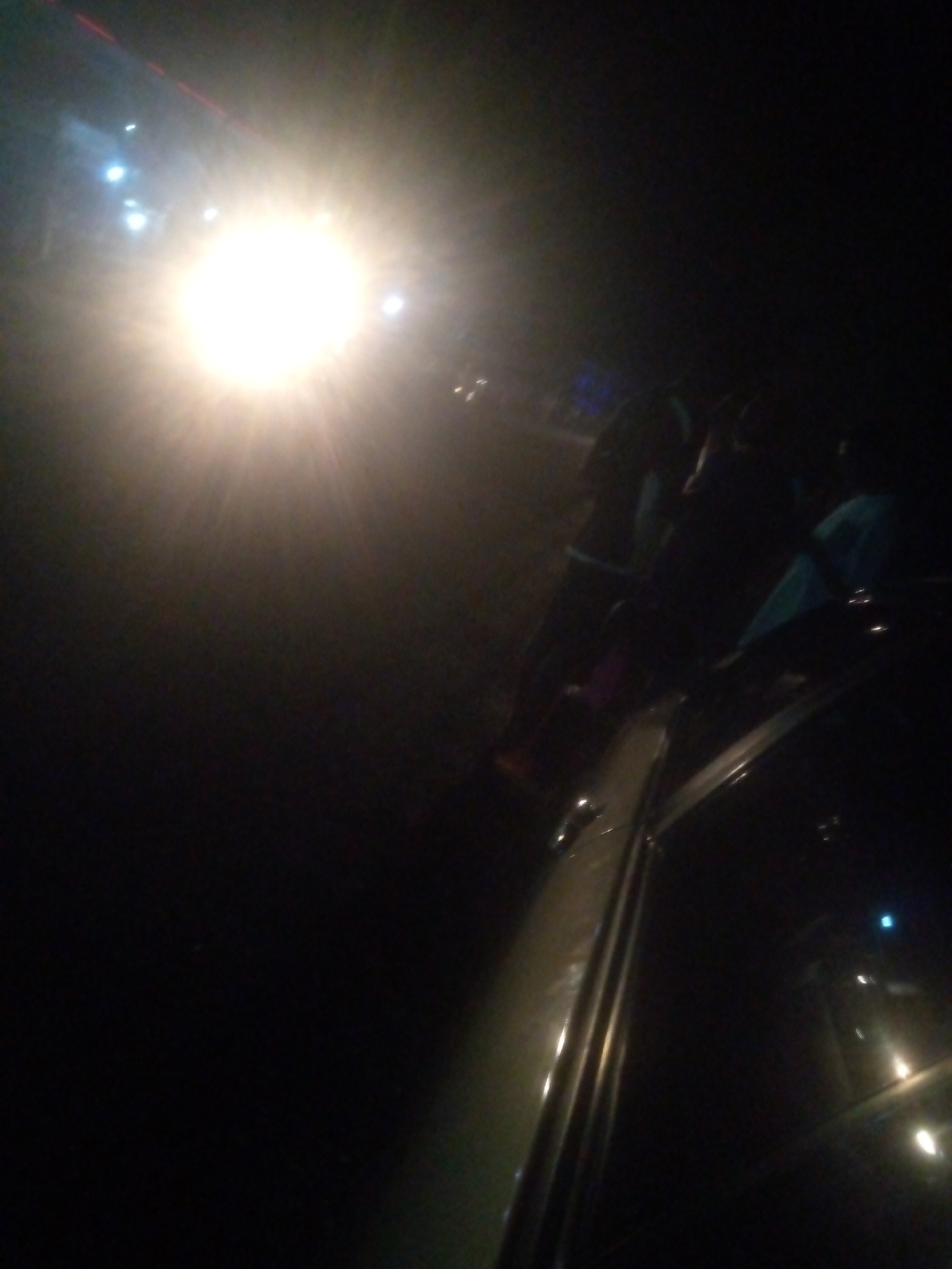 This image depicts a nighttime scene with a prominent, bright light source originating from the upper-left quadrant, casting significant lens flare across the upper-left and central portions of the frame. The light illuminates a hazy atmosphere.
In the middle-right to upper-right section, several indistinct, silhouetted human figures are visible. One figure on the far right appears to be wearing a light blue or teal garment. Their actions and specific interactions are not discernible due to the darkness and lack of detail.
The right side of the image features a segment of a vehicle, likely a car, seen from an oblique angle. A light-colored body panel or roofline runs diagonally across this section. Part of a car door handle and window frame are visible in the lower-middle-right. Reflections of distant lights are apparent on the vehicle's surface and in the far-right background.
The overall environment is dark, suggesting an outdoor setting at night, possibly a street or roadside. There are no specific visual cues within the image itself to confirm the location as Jalingo, Nigeria, other than the context provided.