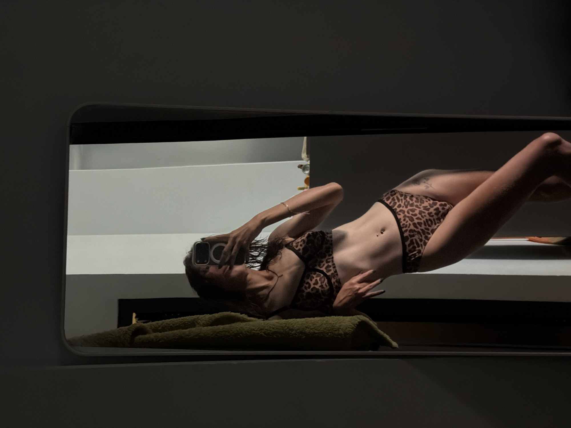 The image is a reflection of a woman taking a selfie in a mirror. The woman is wearing a leopard print bikini. She is lying on a green towel on a surface, possibly a bathroom counter. The background features a white wall and a black surface below. The scene is dimly lit. The image's location is unspecified, and no indication of being in Tay Nha Trang Ward, Vietnam, is present.