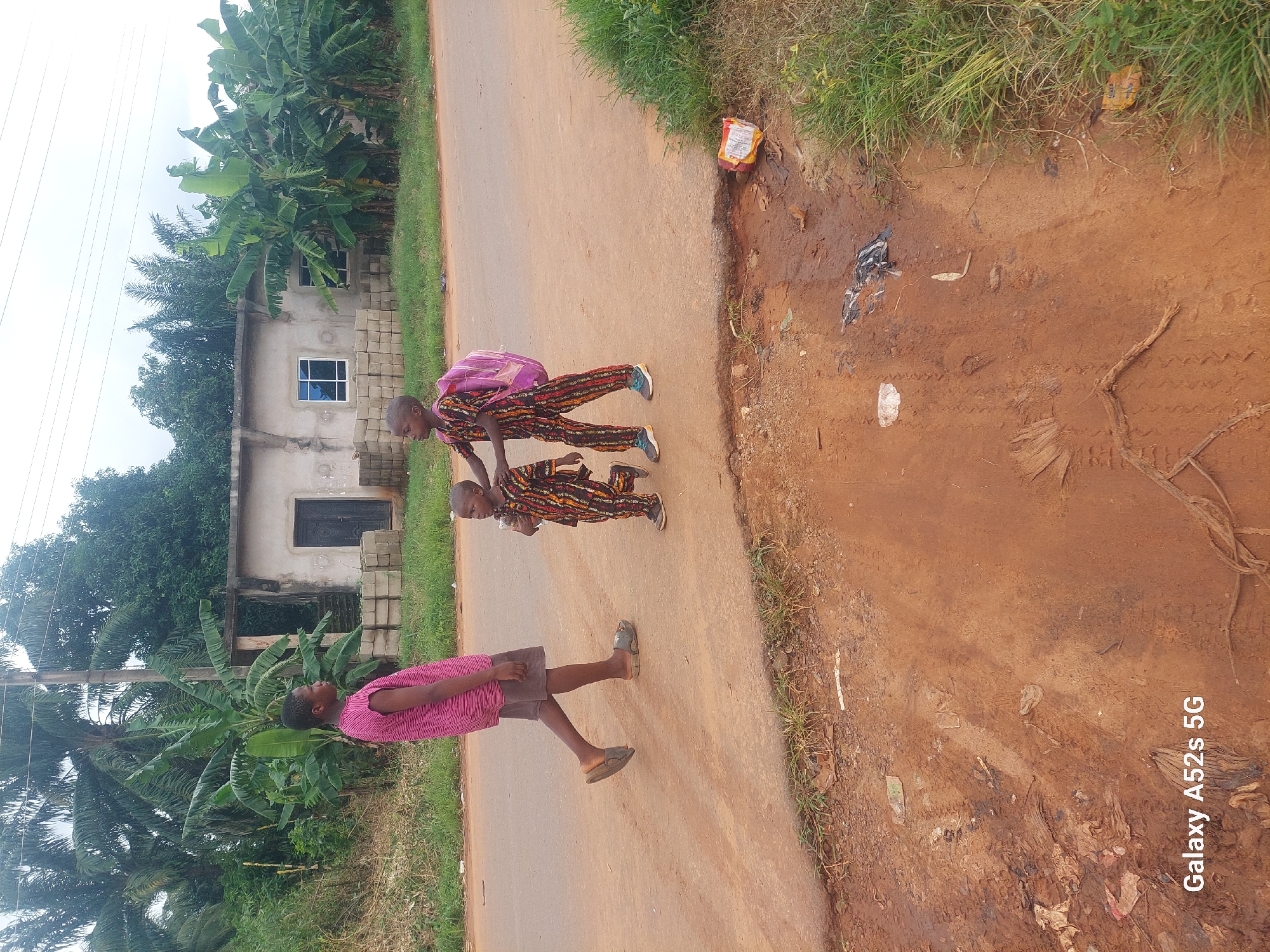 Three children are walking on a dirt road in Umudioka, Nigeria. The child on the left is wearing a pink and black striped shirt and dark shorts. The child in the middle is wearing a colorful patterned outfit and carrying a pink backpack. The child on the right is wearing a similar colorful patterned outfit. In the background, there is a partially constructed building with windows and a doorway. Banana trees and other foliage surround the area. The road appears to be made of reddish-brown dirt, with some grass and debris visible along the edges. A single power line extends across the top of the image.