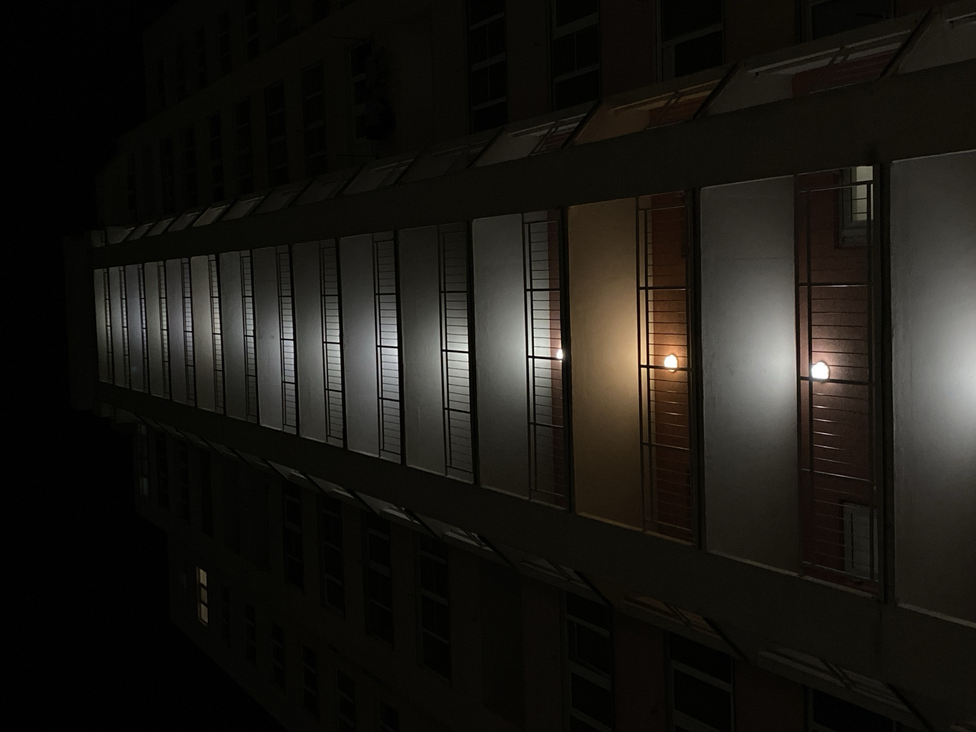 The image displays a multi-story building at night, captured from a low, upward-angled perspective. A prominent architectural element, likely a long corridor or balcony, stretches horizontally across the mid-ground. This structure features a series of rectangular sections, each fronted by frosted or translucent panels. Behind these panels, vertical metal grilles are visible.
Within several of these sections, interior lights are illuminated, casting a soft glow. Some lights emit a warm, orange-yellow hue, while others project a cooler, white light, appearing as distinct luminous spheres. The overall scene suggests a quiet nocturnal environment, with some building occupants having their lights on.
Above and below this main illuminated section, the darker facade of the building is visible, punctuated by numerous windows, most of which are unlit and recede into the profound darkness of the night. No people or specific activities are discernible. The time of day is clearly night. There are no explicit weather indicators, and no visible text. The architectural style hints at a functional building, but its specific location remains unknown.