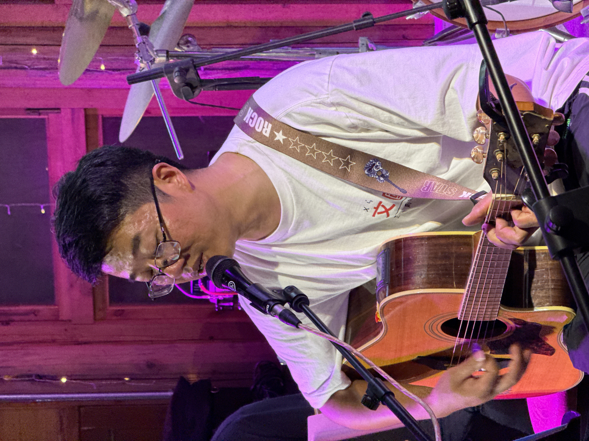 A musician plays an acoustic guitar while singing into a microphone. He wears glasses, a white T-shirt, and a guitar strap labeled "Rock Star". A drum set is partially visible in the background. The scene is illuminated with purple lighting, and a wooden wall is visible behind the musician. It is unlikely one can determine the exact location of this image in the city of Beibei, China.