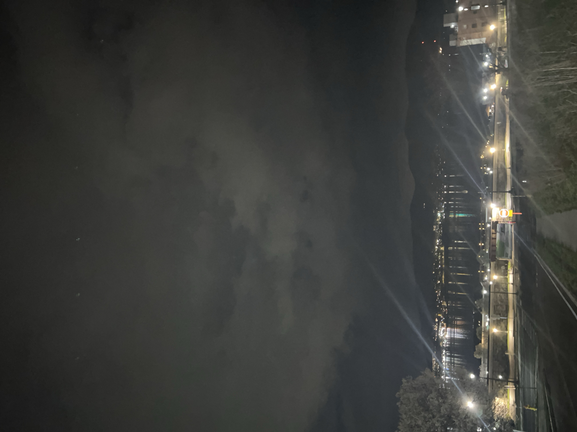 The image shows a night view of the City of Newburgh, United States of America, along the Hudson River. A dark, overcast sky hangs over the river, with reflections of city lights visible on the water's surface. A roadway runs along the shoreline, illuminated by streetlights. Buildings and trees are visible in the background.